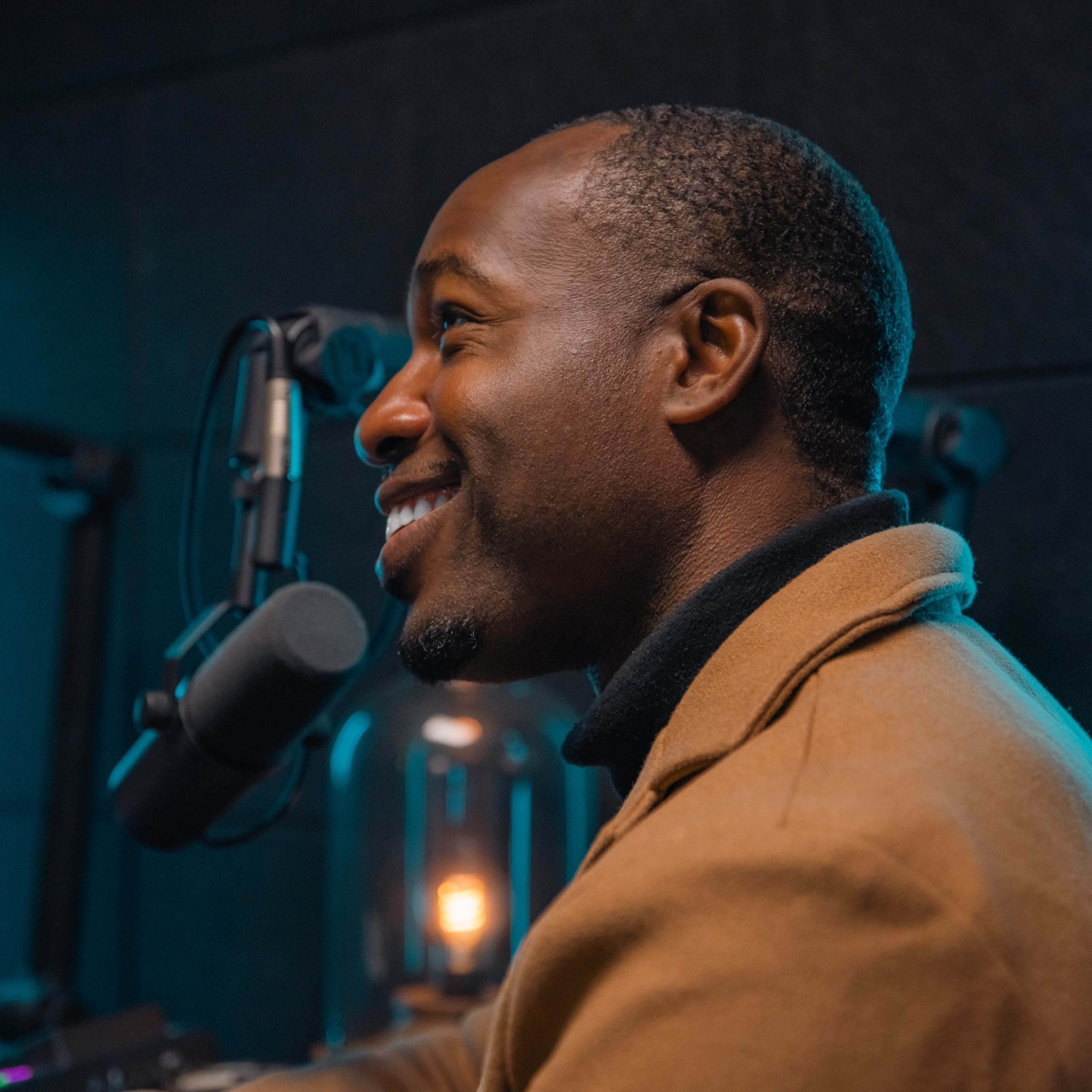 A man is sitting in a recording studio, smiling while speaking into a microphone, with blue neon lights illuminating the background.