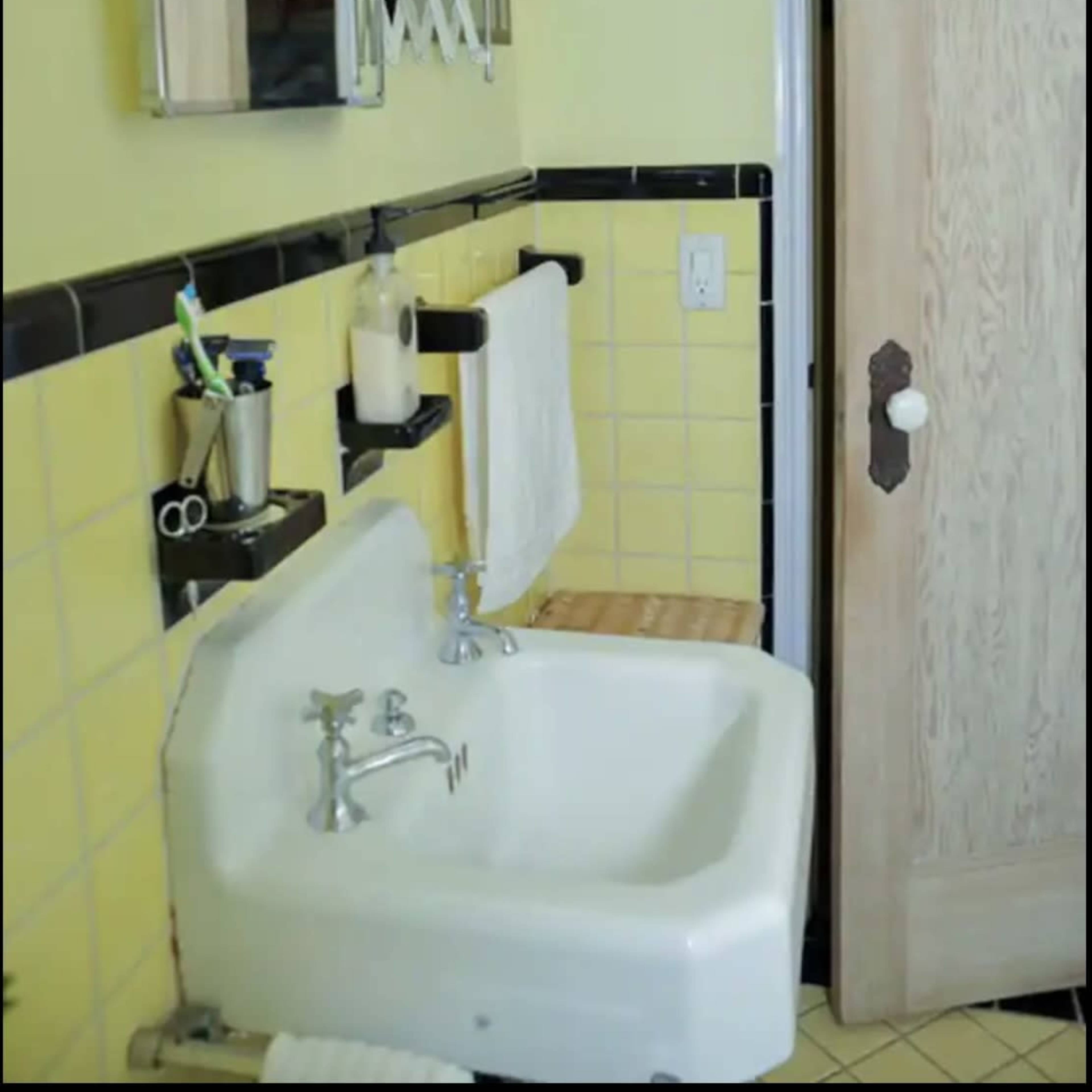 A small bathroom with yellow tiled walls, a white sink mounted on a wall, and a wooden shelf, with a partially open door on the right.