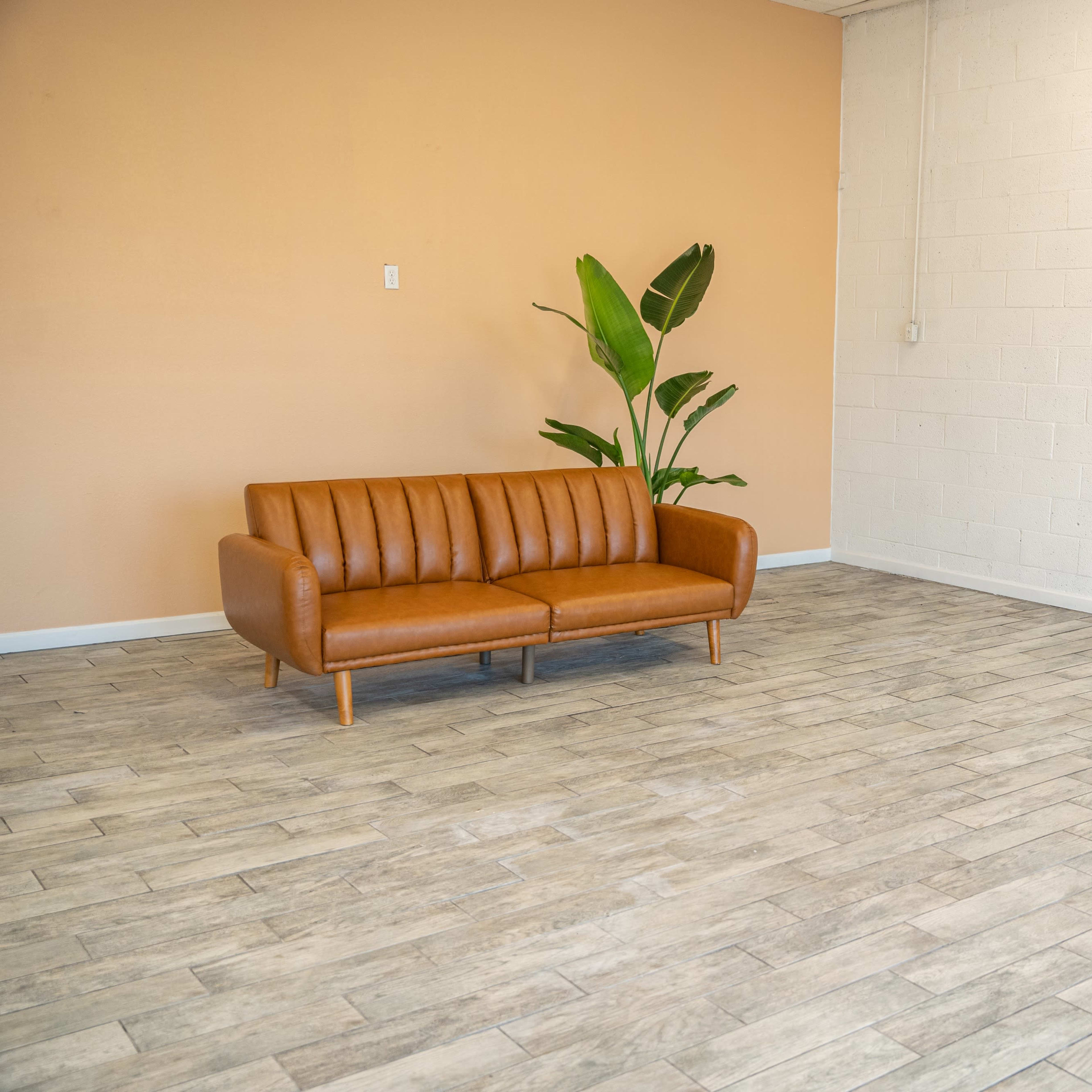 A spacious room with a brown leather couch and a potted plant against an orange wall and a white brick wall.