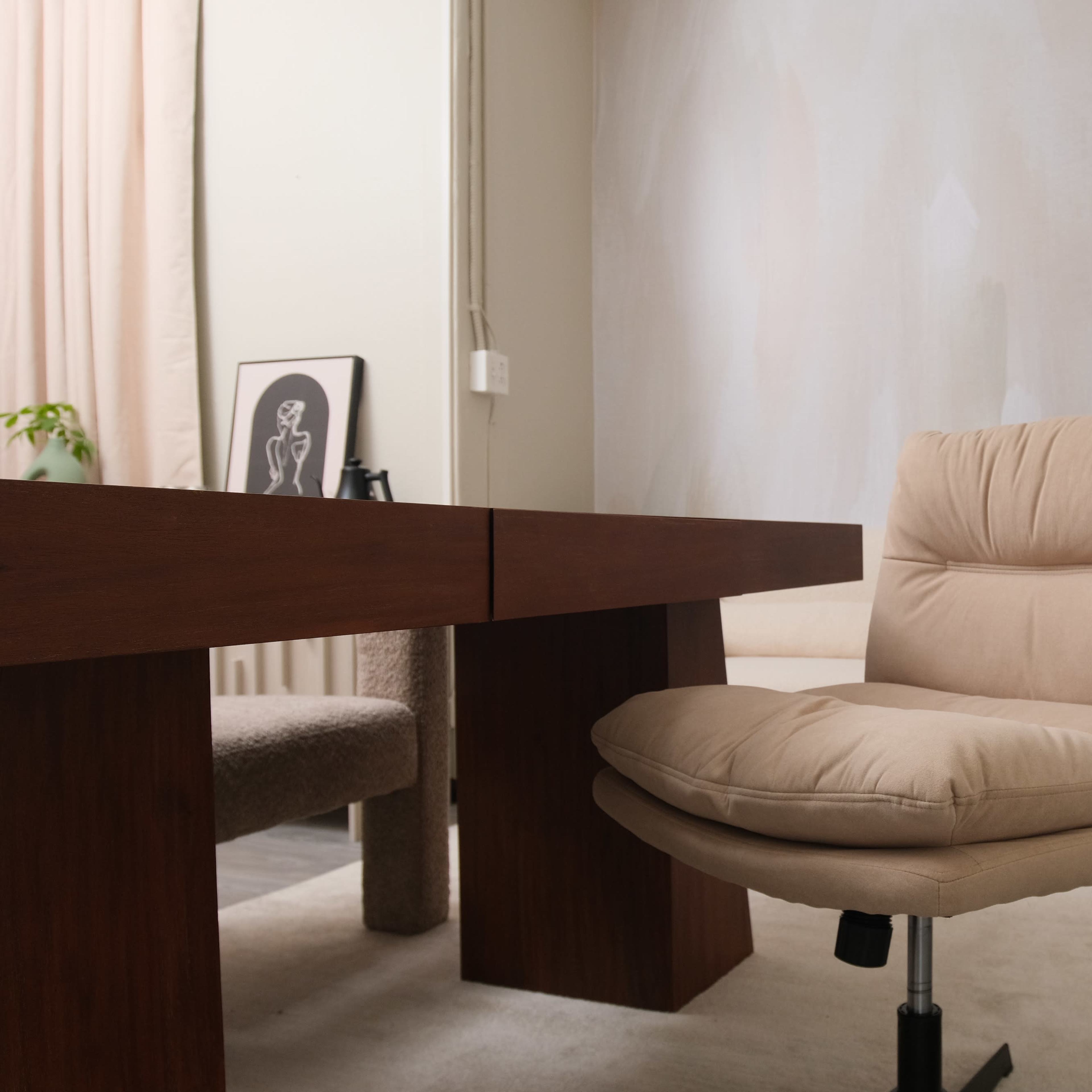 A minimalist office setting featuring a wooden desk, a beige rolling chair, and a potted plant in the corner.
