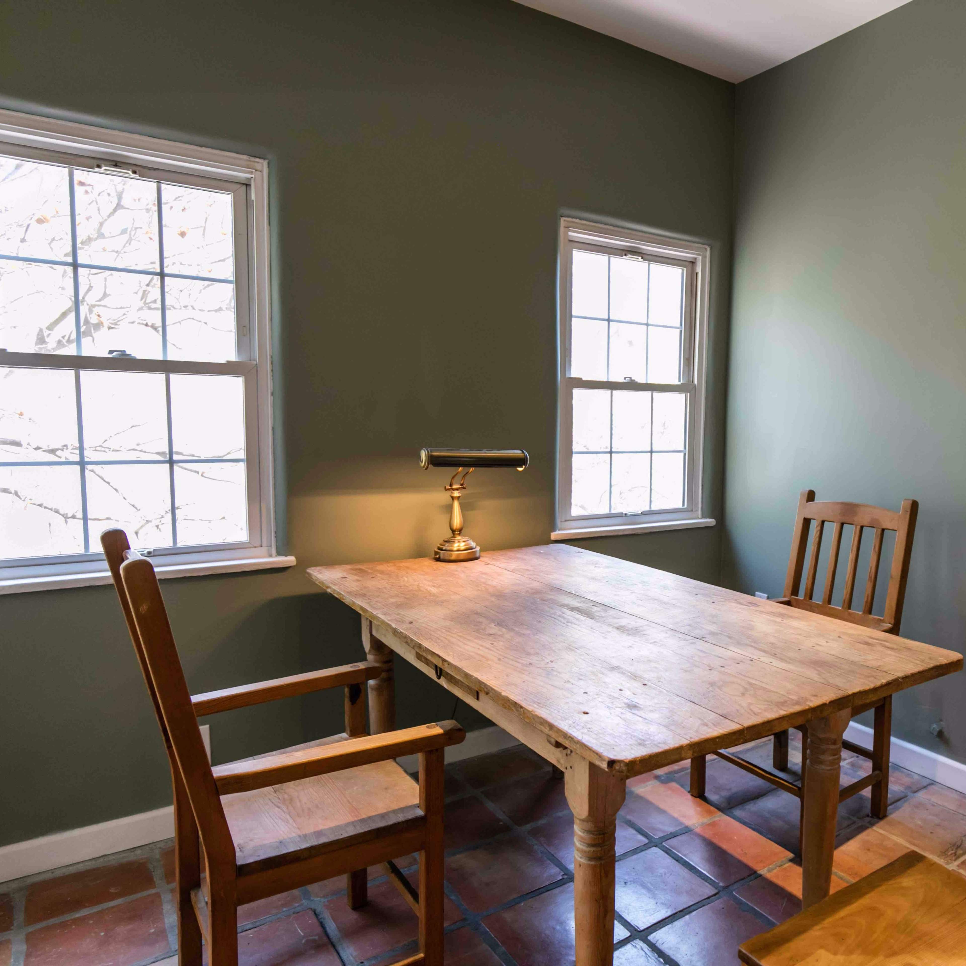 A wooden table with a lamp is positioned between two wooden chairs in a green-walled room with two windows.