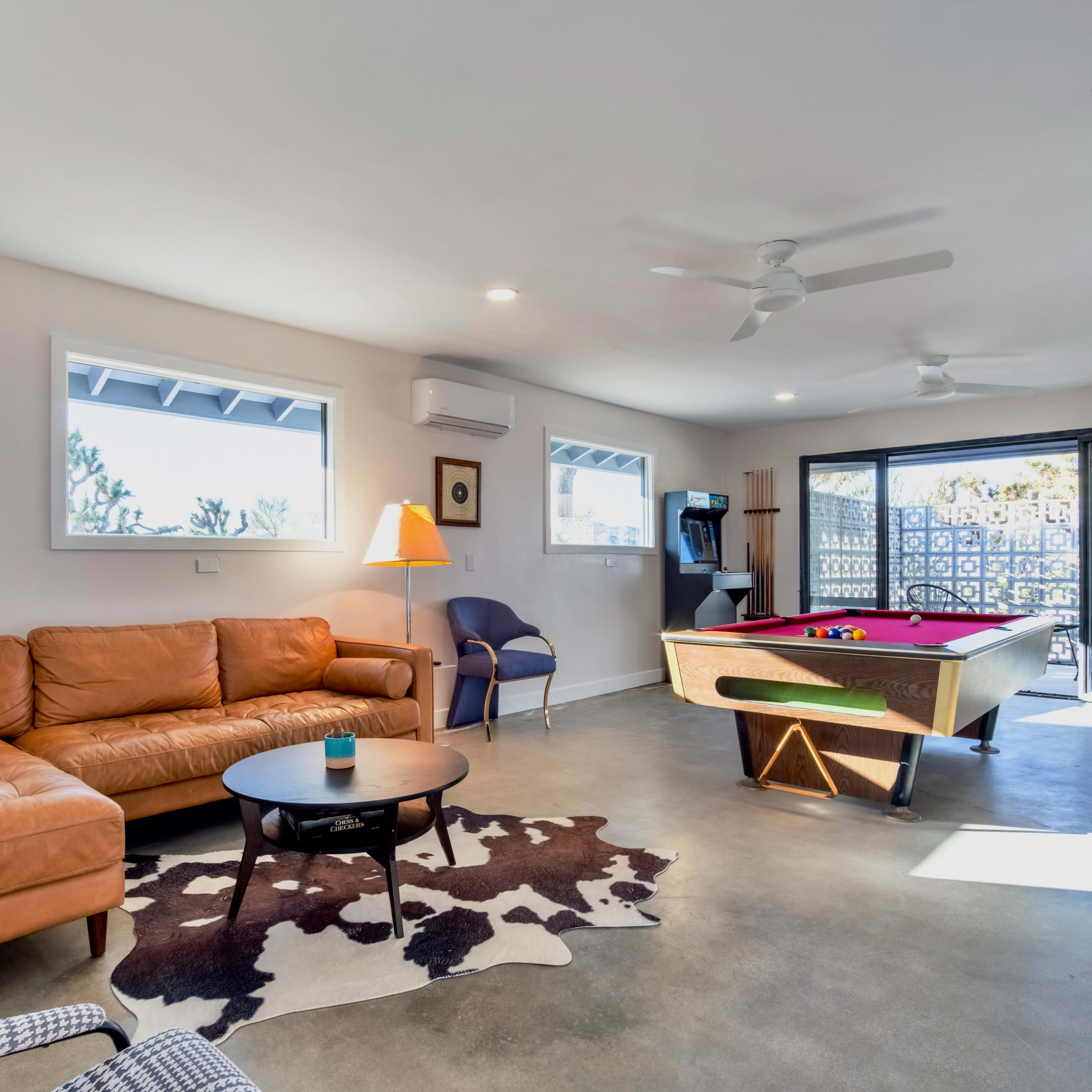 A spacious living room features a tan leather sofa, a round coffee table, and a pool table, with large windows allowing natural light to enter.