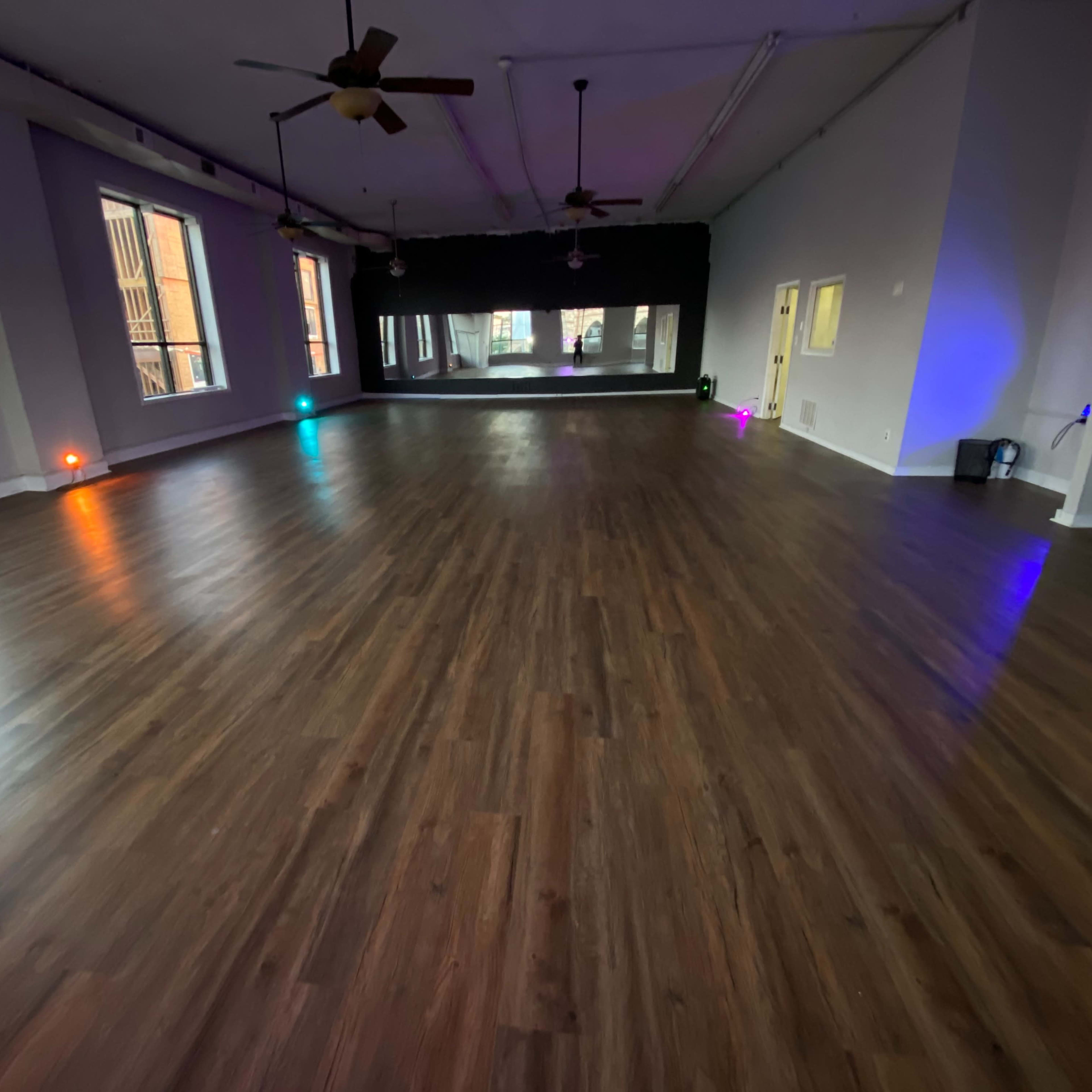 A spacious, empty room with a wooden floor, large windows, and colored lights at the corners.