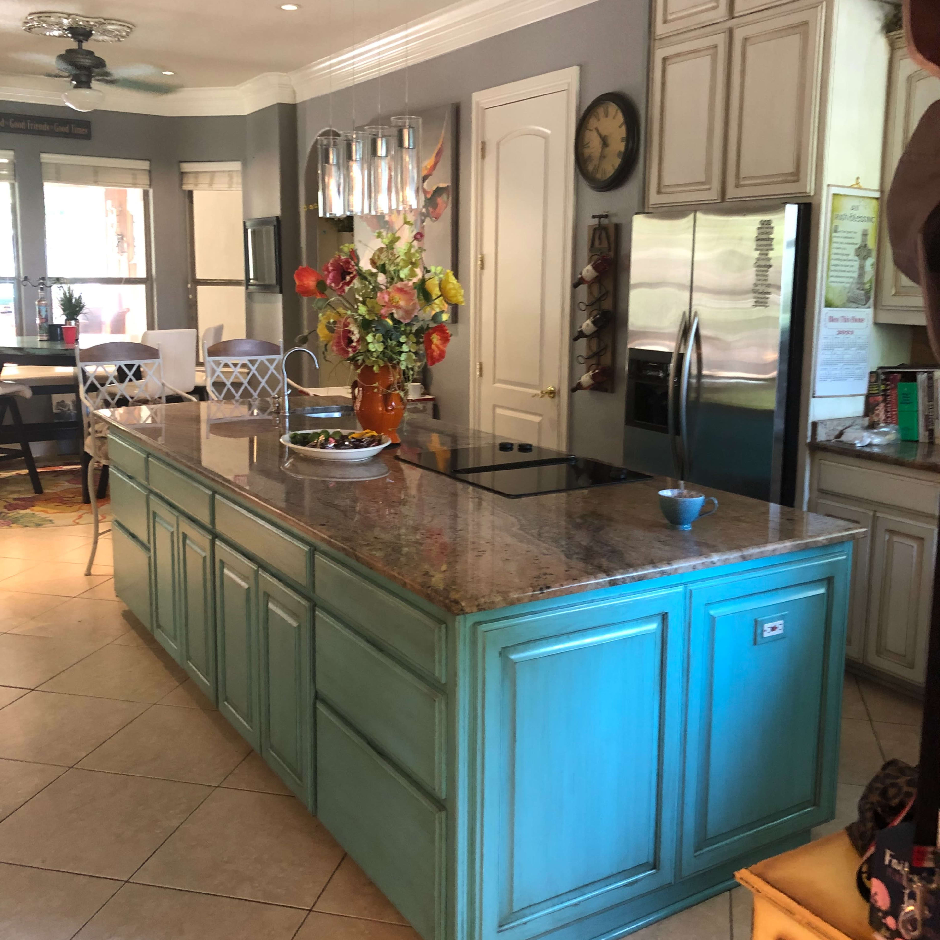 The kitchen features an island with a bright turquoise finish, a granite countertop, and various appliances, surrounded by light-colored cabinetry and a dining area in the background.