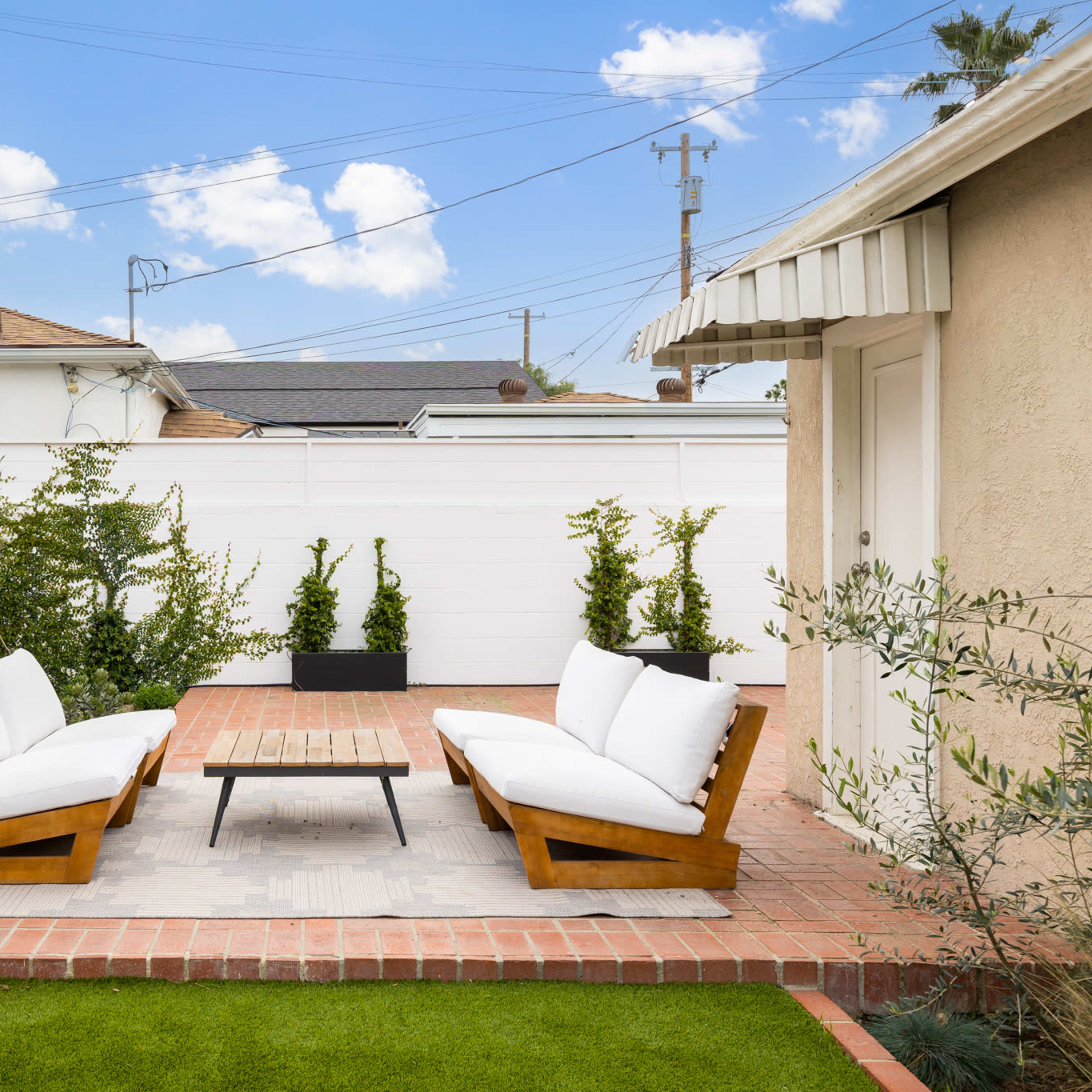 A cozy outdoor seating area features two wooden benches with white cushions arranged around a rectangular coffee table on a brick patio, surrounded by greenery and a white fence.