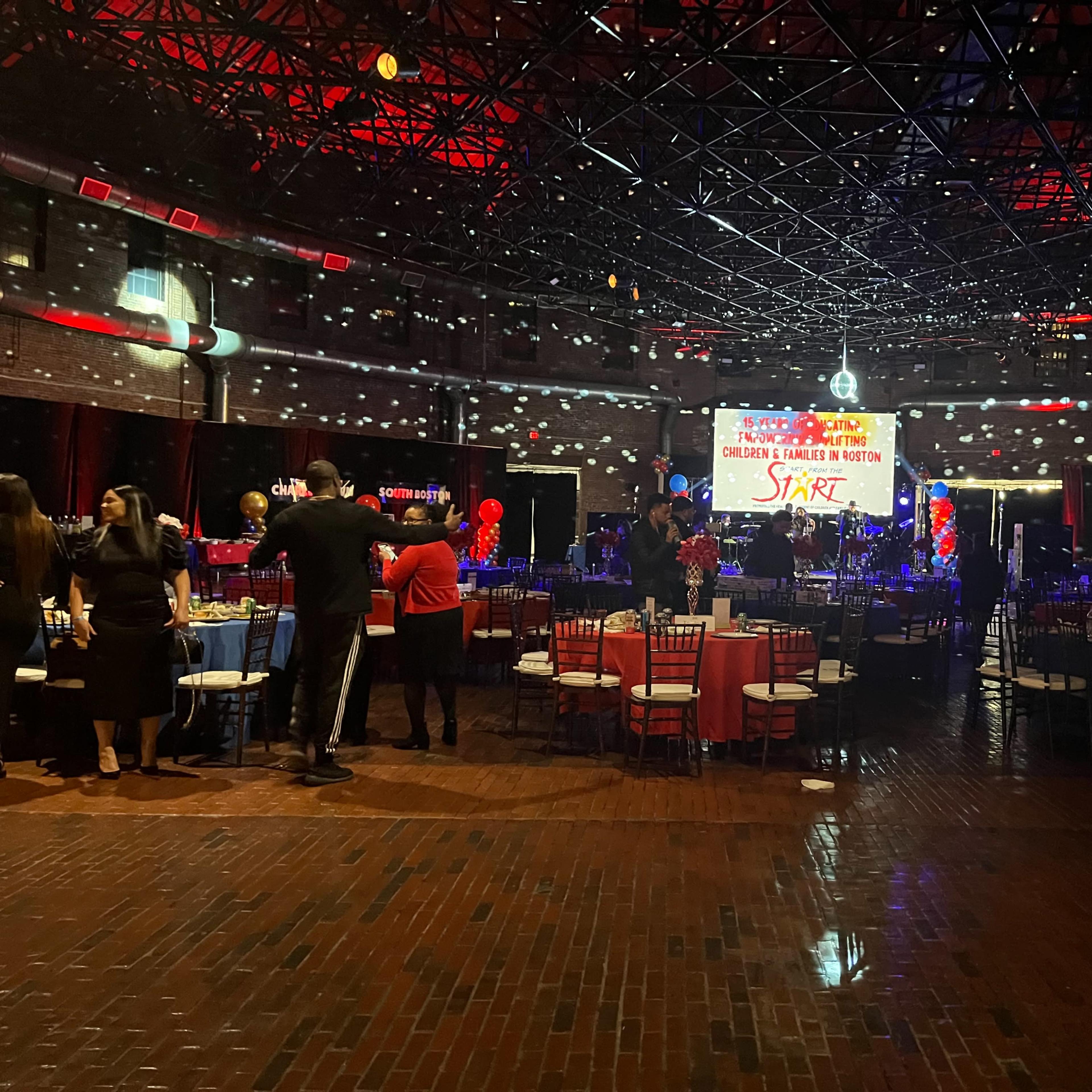 A spacious event hall features decorated tables, colorful lighting, and a large banner that reads "Start" in the background.