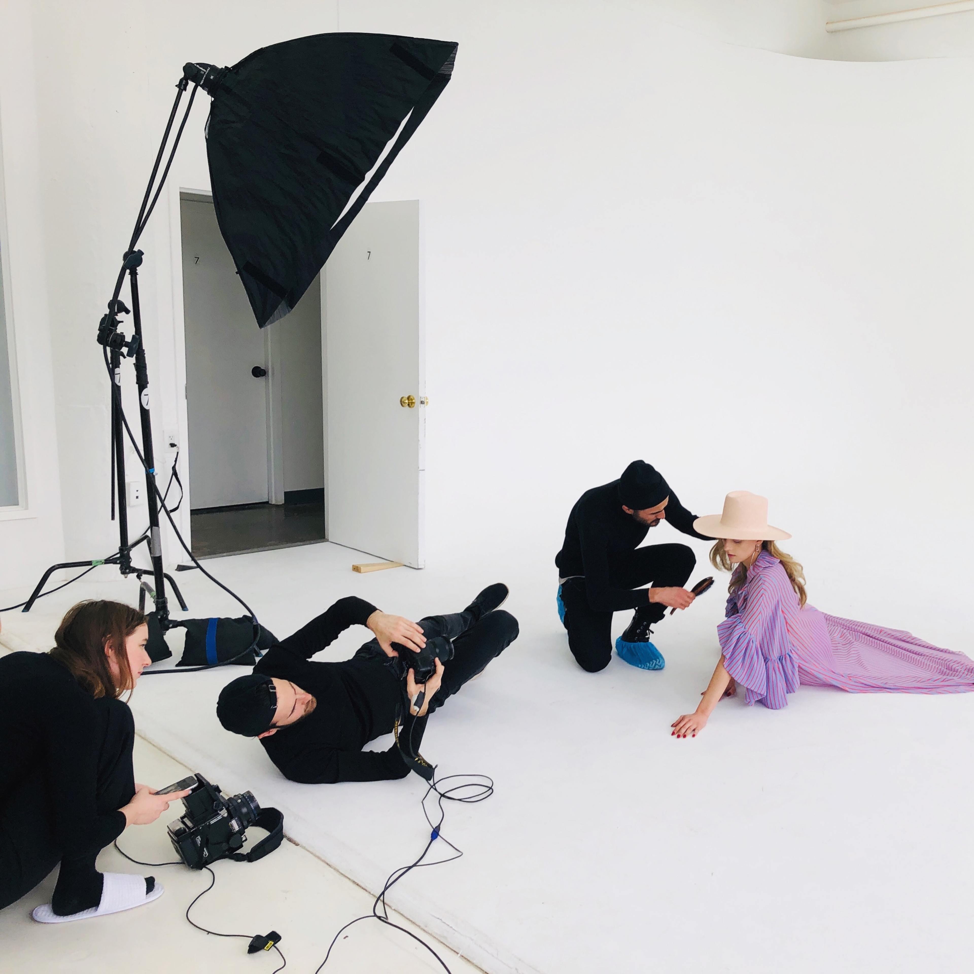A photography crew is capturing a fashion shoot featuring three models in a white studio, with one photographer lying on the ground and two crew members adjusting lighting equipment.