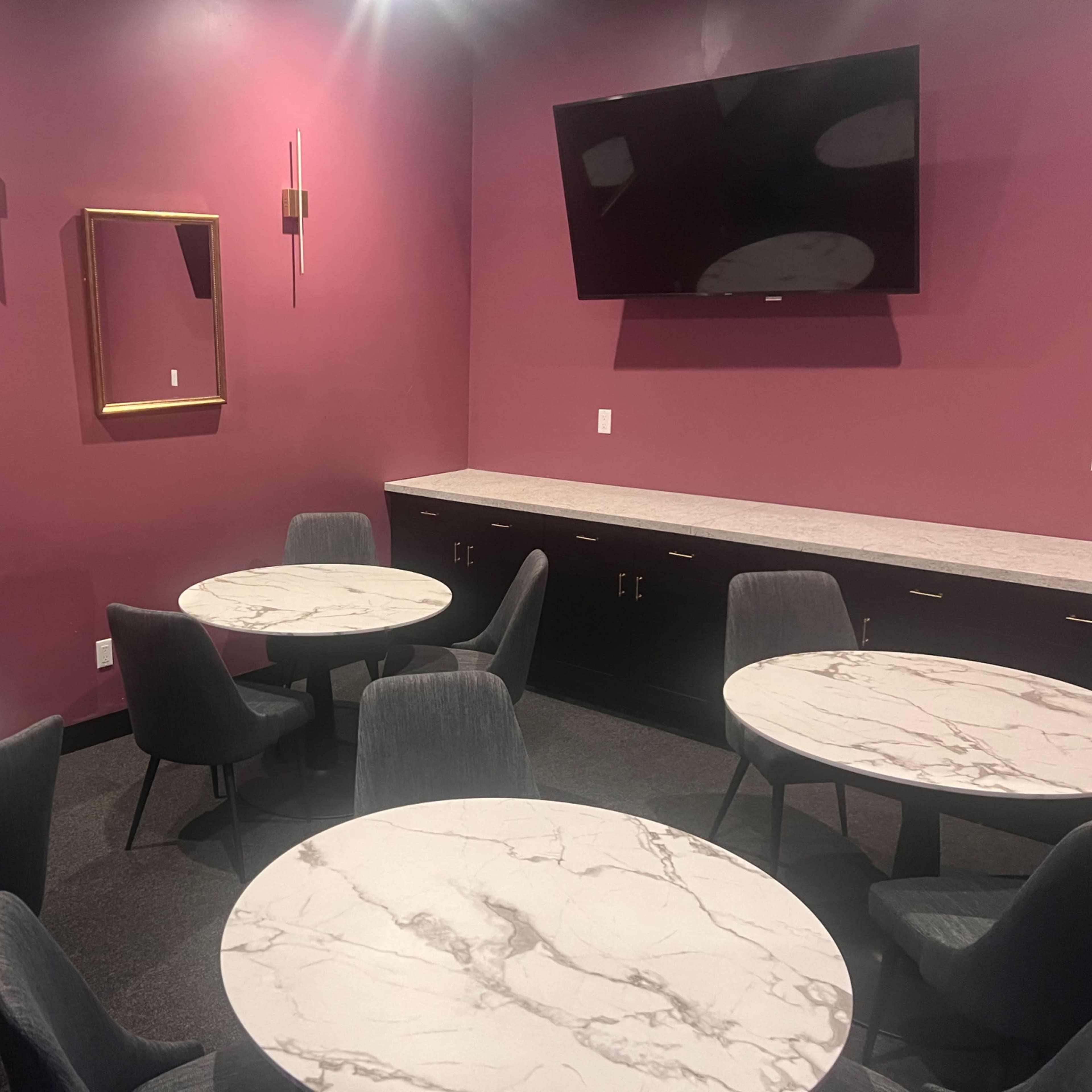 A small, modern dining area with round marble tables and black chairs against a deep pink wall, featuring a mounted television and a mirror.