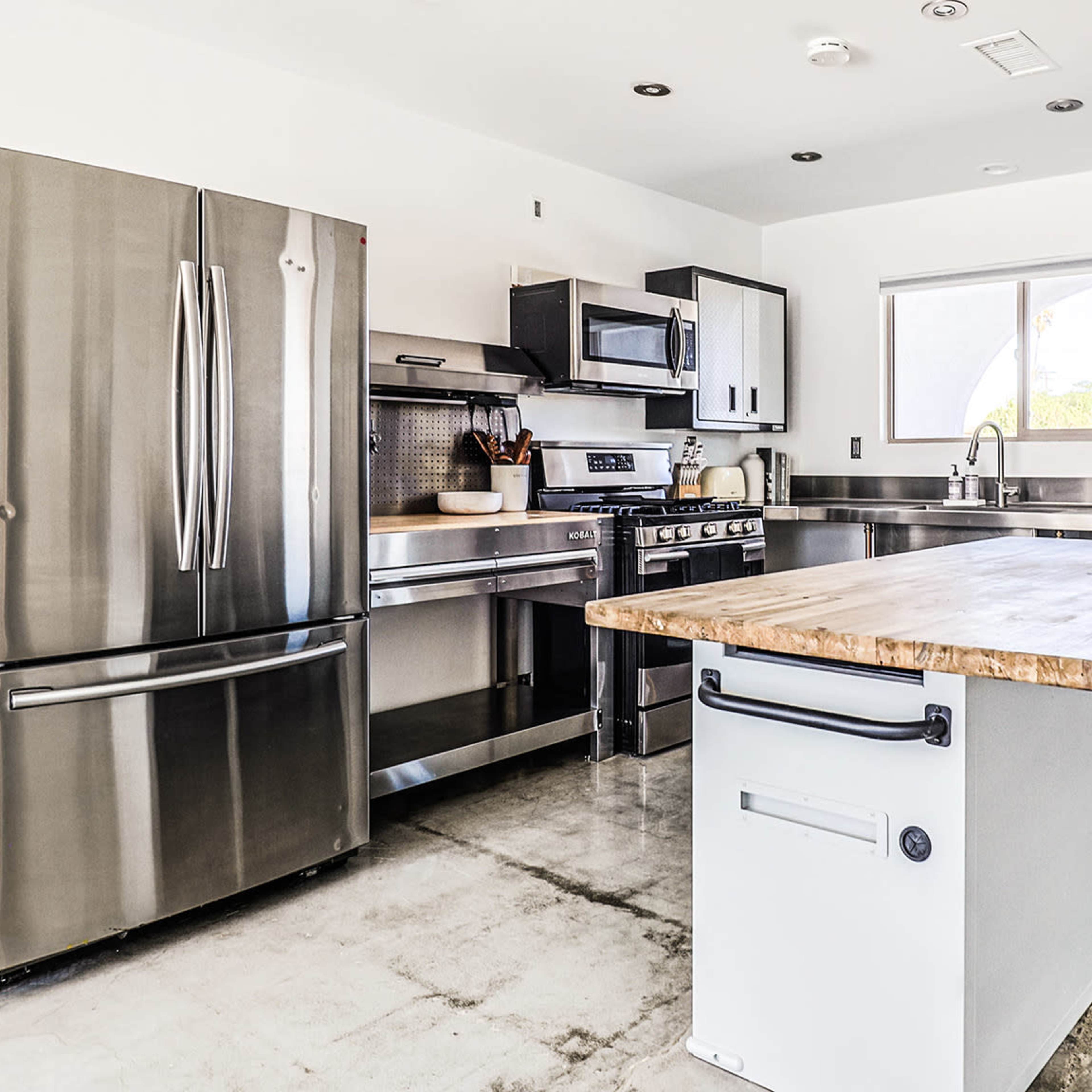 A modern kitchen featuring stainless steel appliances, a central wooden island, and minimalist cabinetry against a white wall.