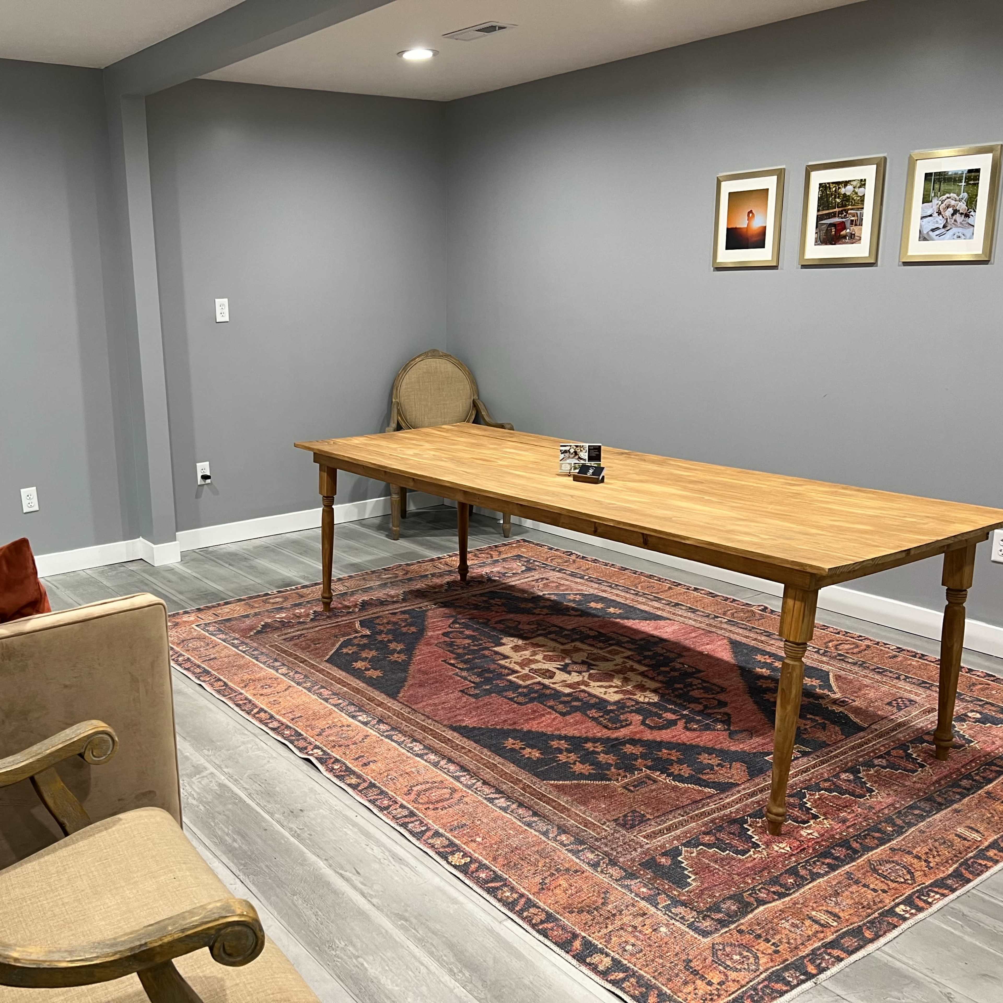 A simple interior room with a wooden table, two chairs, a couch with cushions, and framed pictures on the gray wall.