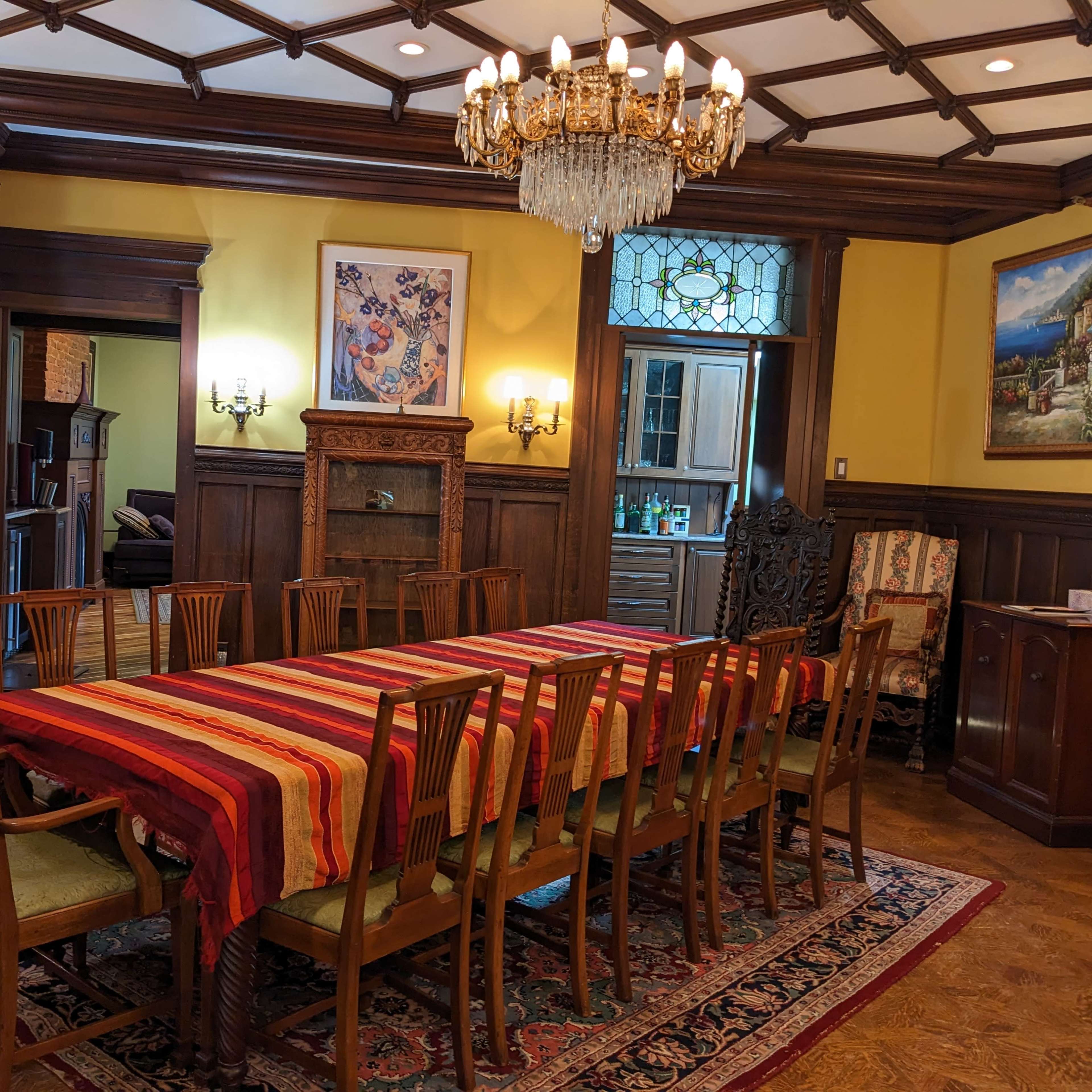 A large dining room featuring a long table set with a striped tablecloth, surrounded by wooden chairs, and adorned with a chandelier and framed artwork on the walls.