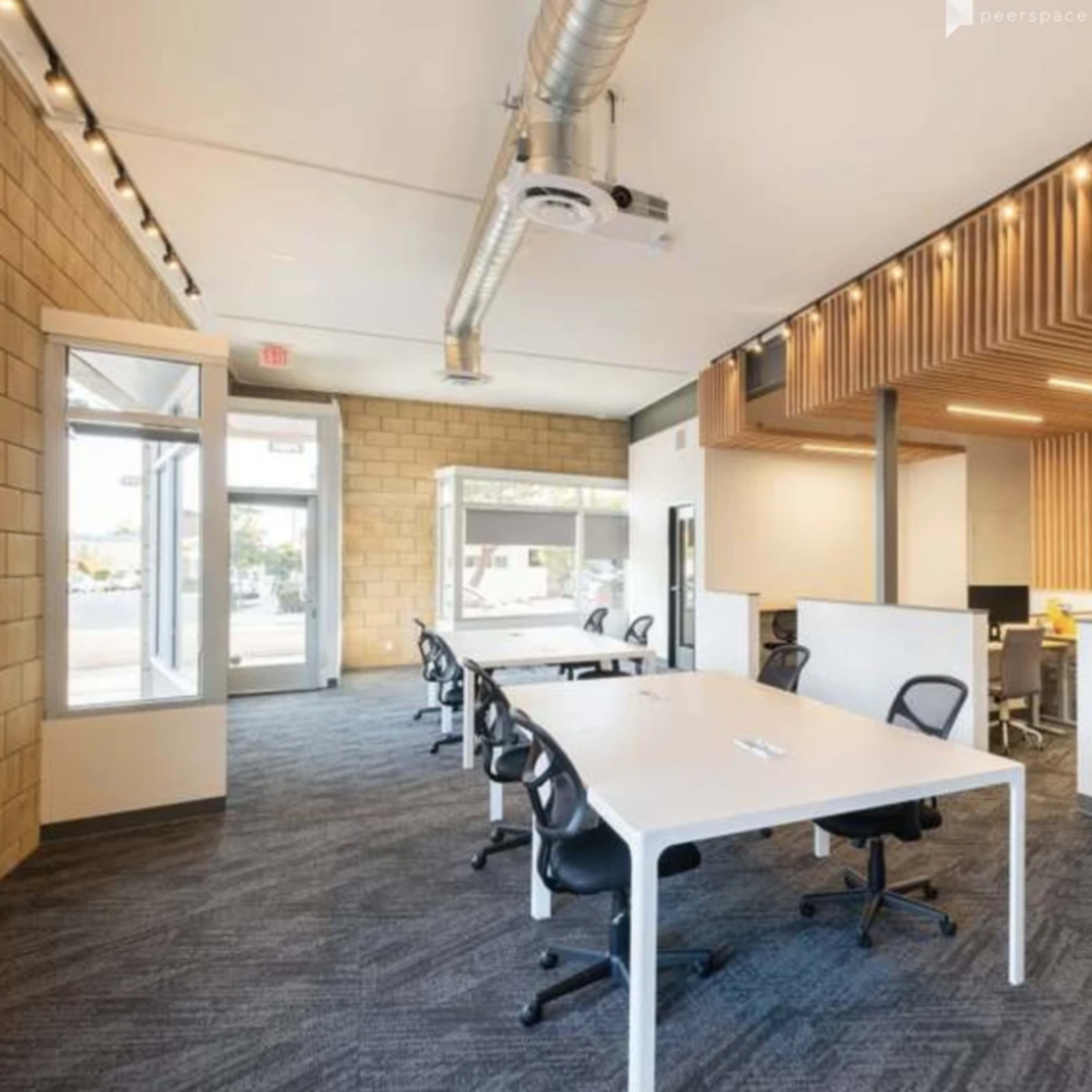 A modern office space with several white desks and black chairs, large windows, and a wood-paneled ceiling.