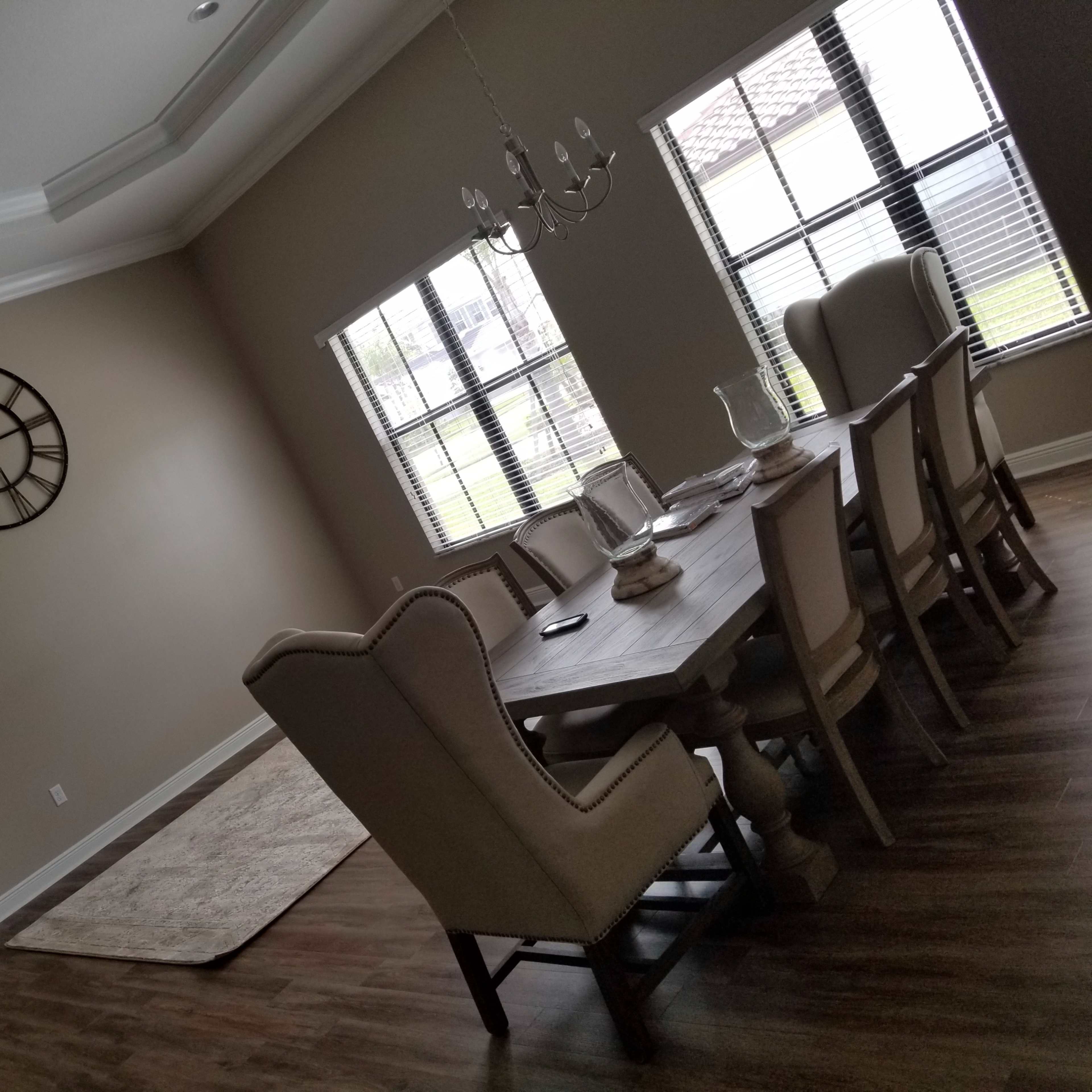 A dining room features a large table surrounded by upholstered chairs, with a clock on the wall and windows allowing natural light.