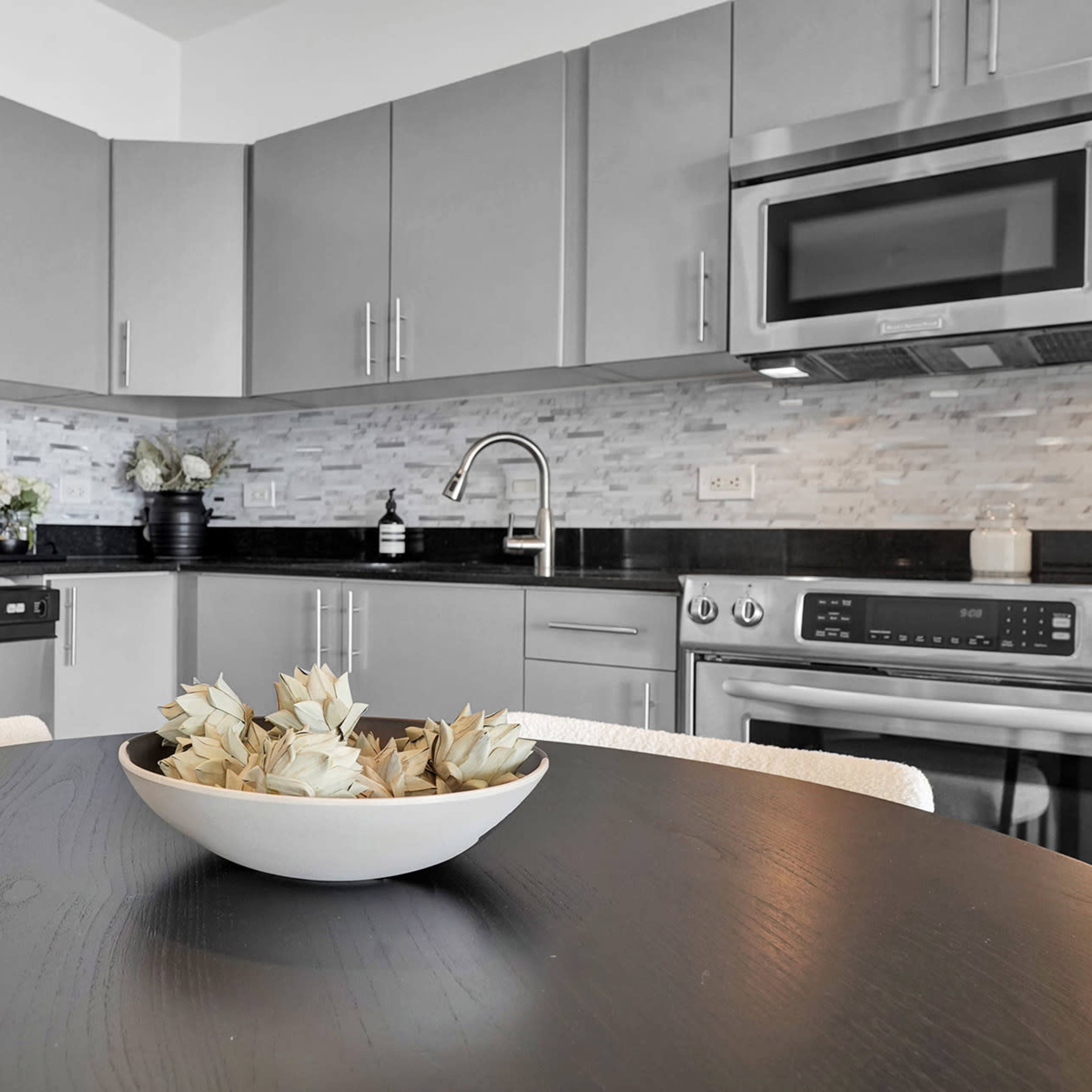 A modern kitchen features stainless steel appliances, gray cabinetry, and a black countertop with a bowl of decorative items on the table.