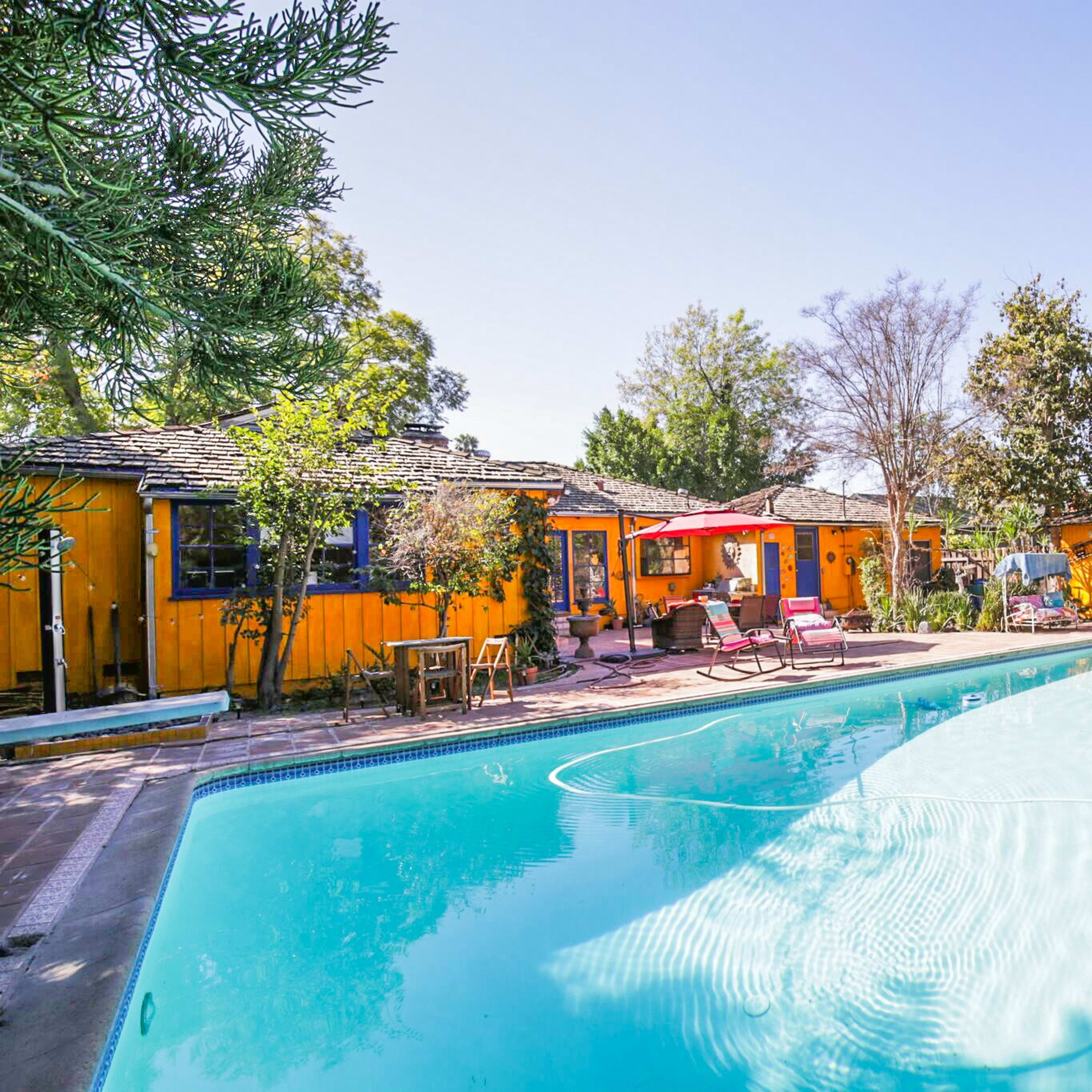A clear swimming pool with a surrounding patio features colorful cabins and lush greenery.