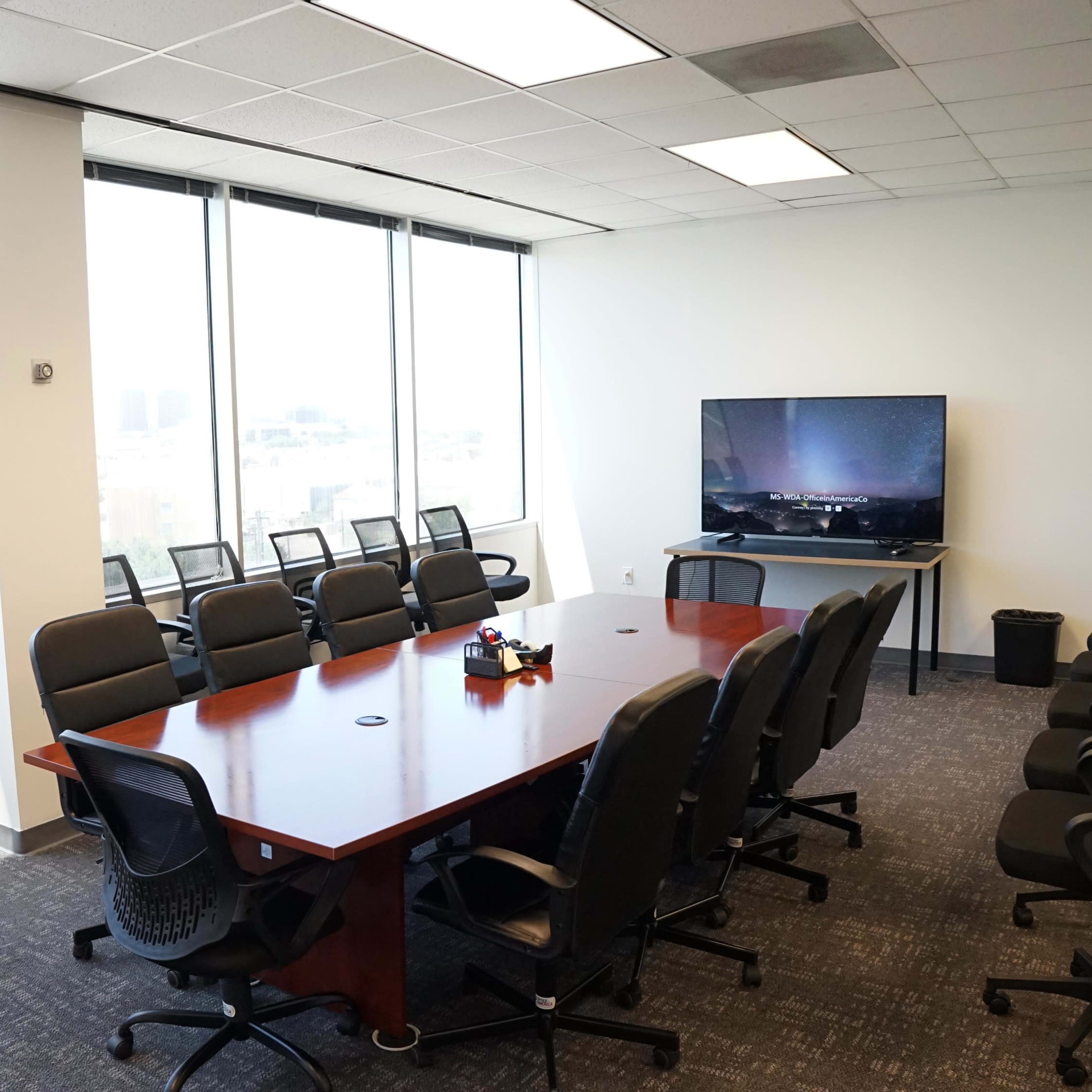 A spacious conference room with a large wooden table and black chairs, overlooking a cityscape through floor-to-ceiling windows.