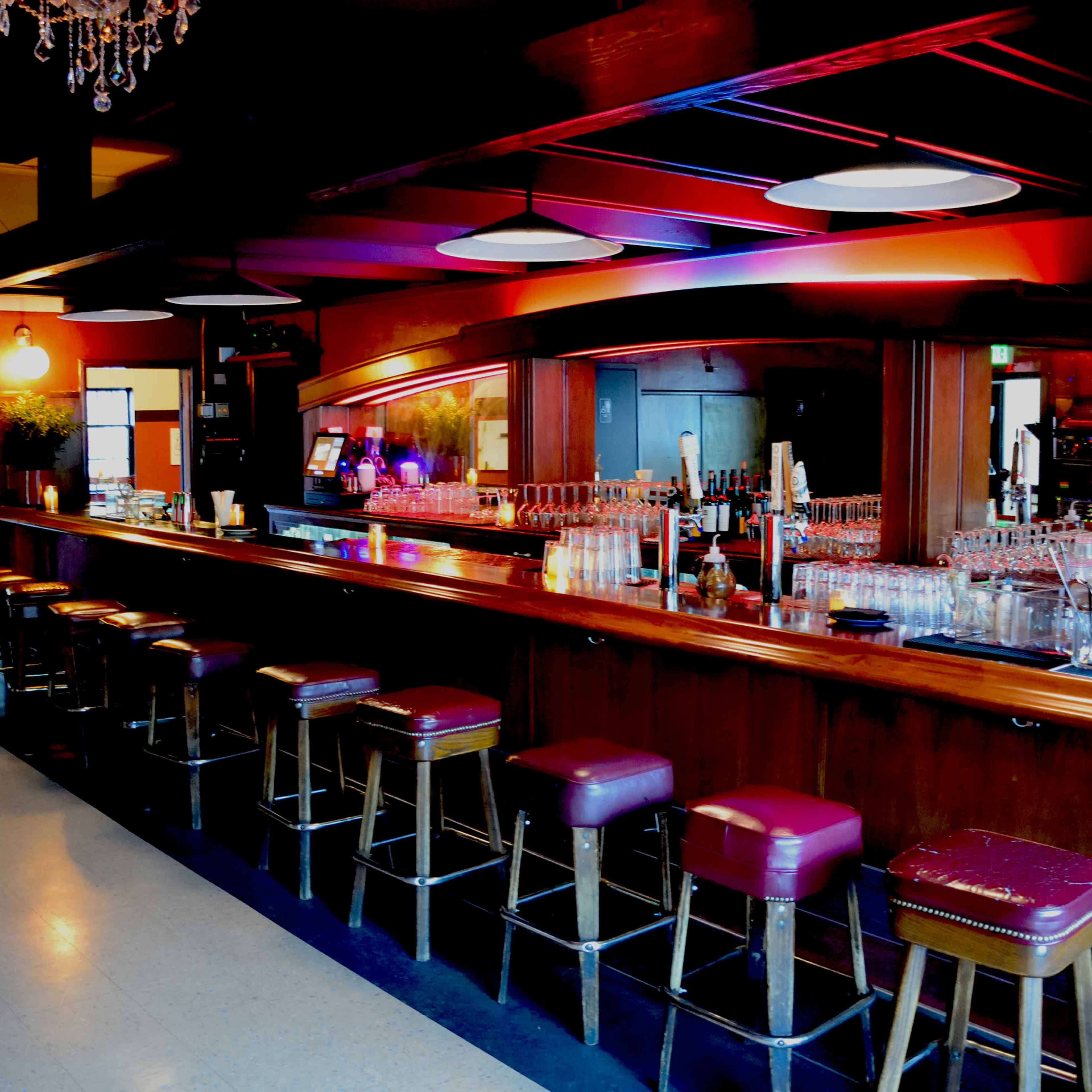 A dimly lit bar with a long wooden counter, several bar stools, and a mirrored back displaying various glassware.