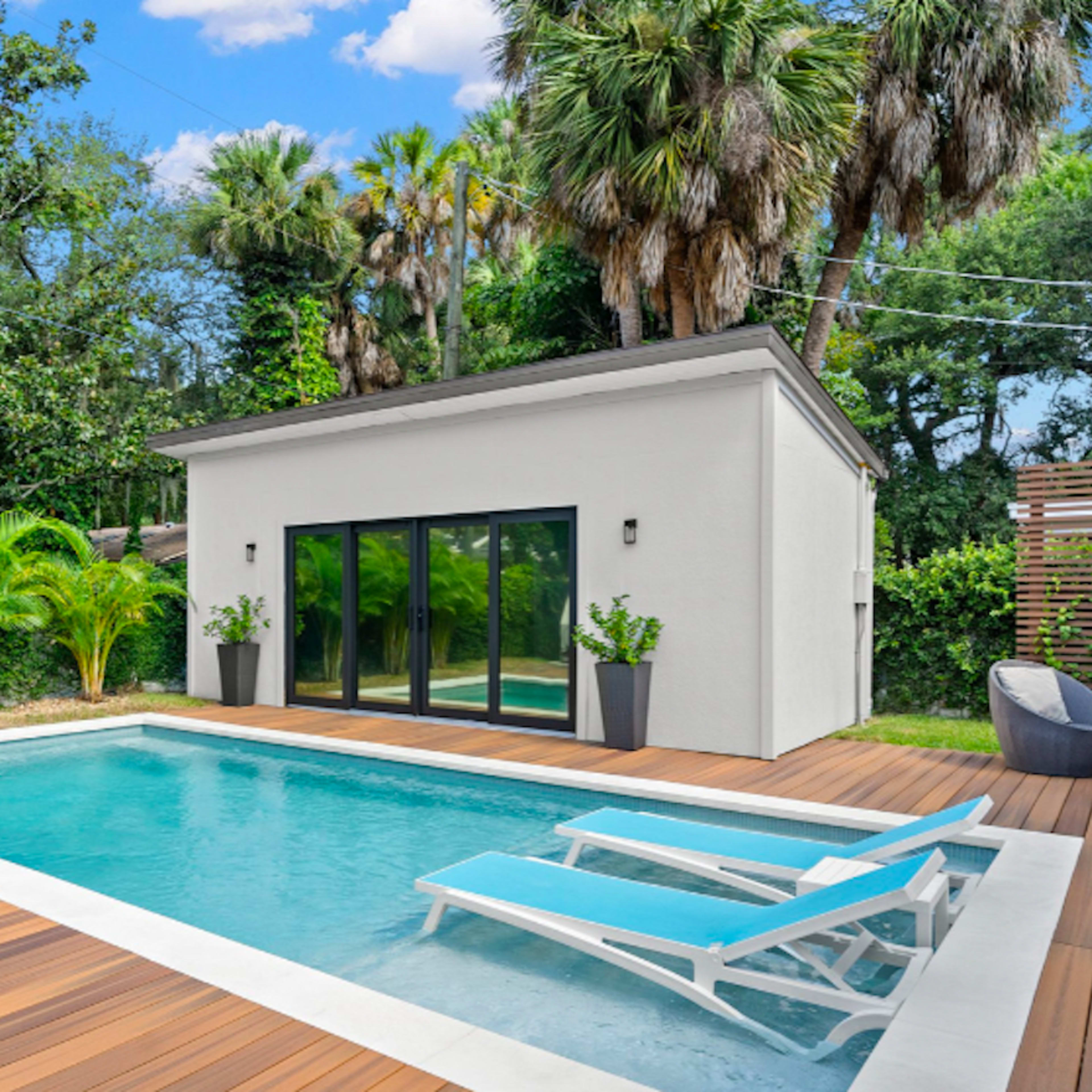 A small pool surrounded by wooden decking, with lounge chairs and a modern building in a lush, tropical garden.