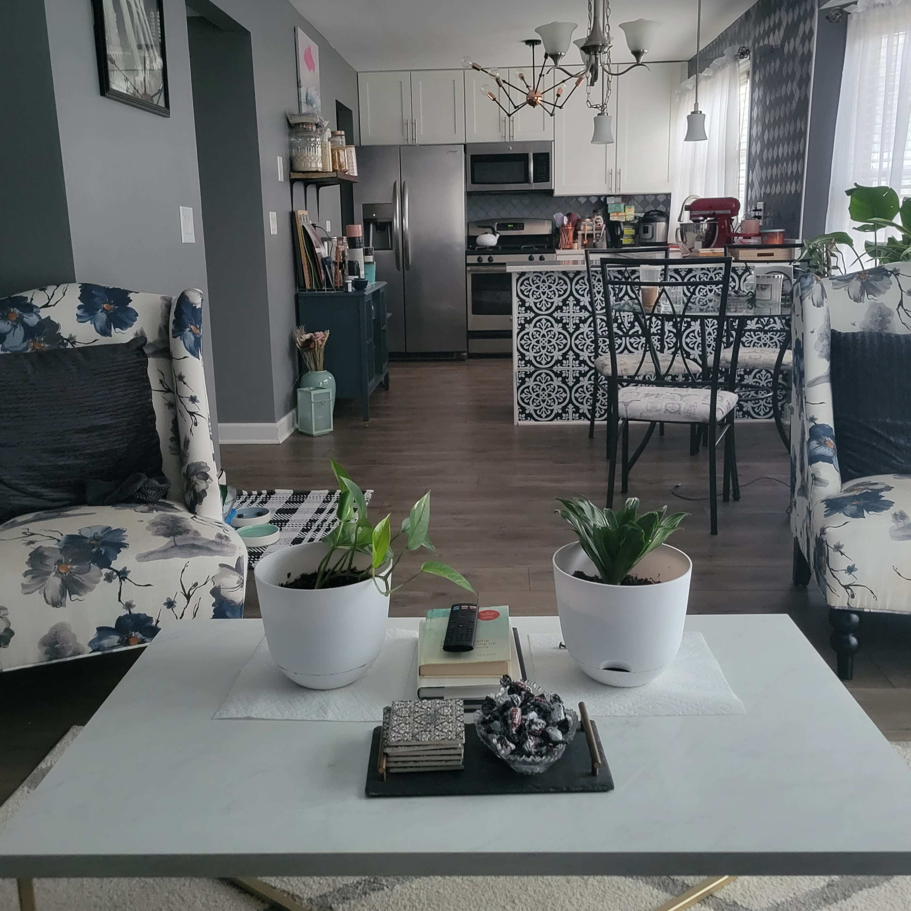 A modern living room with floral-patterned chairs, a coffee table with plants and books, and an open kitchen in the background.