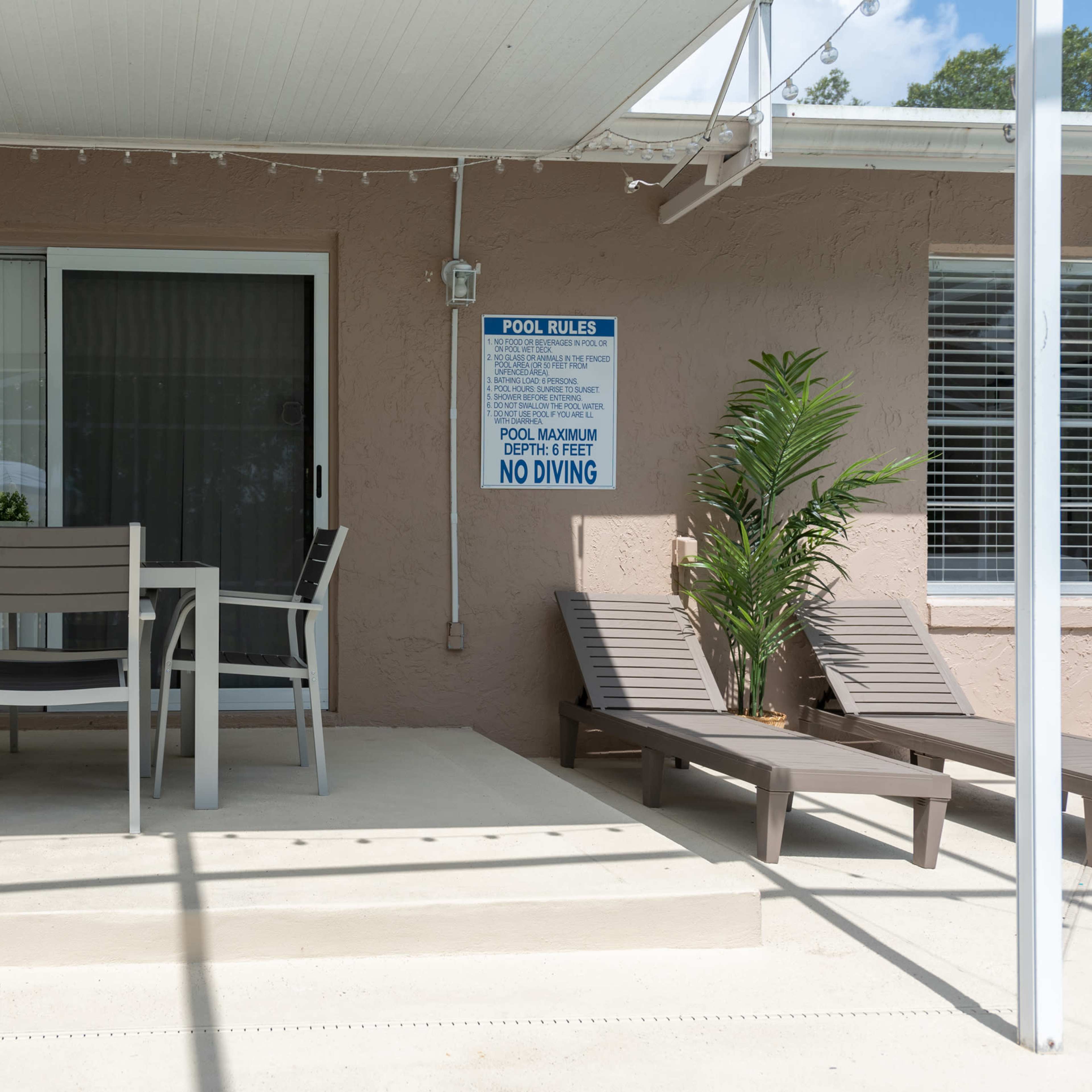 A patio area with a dining table and chairs, a sign about pool rules, and lounging chairs under a covered outdoor space.