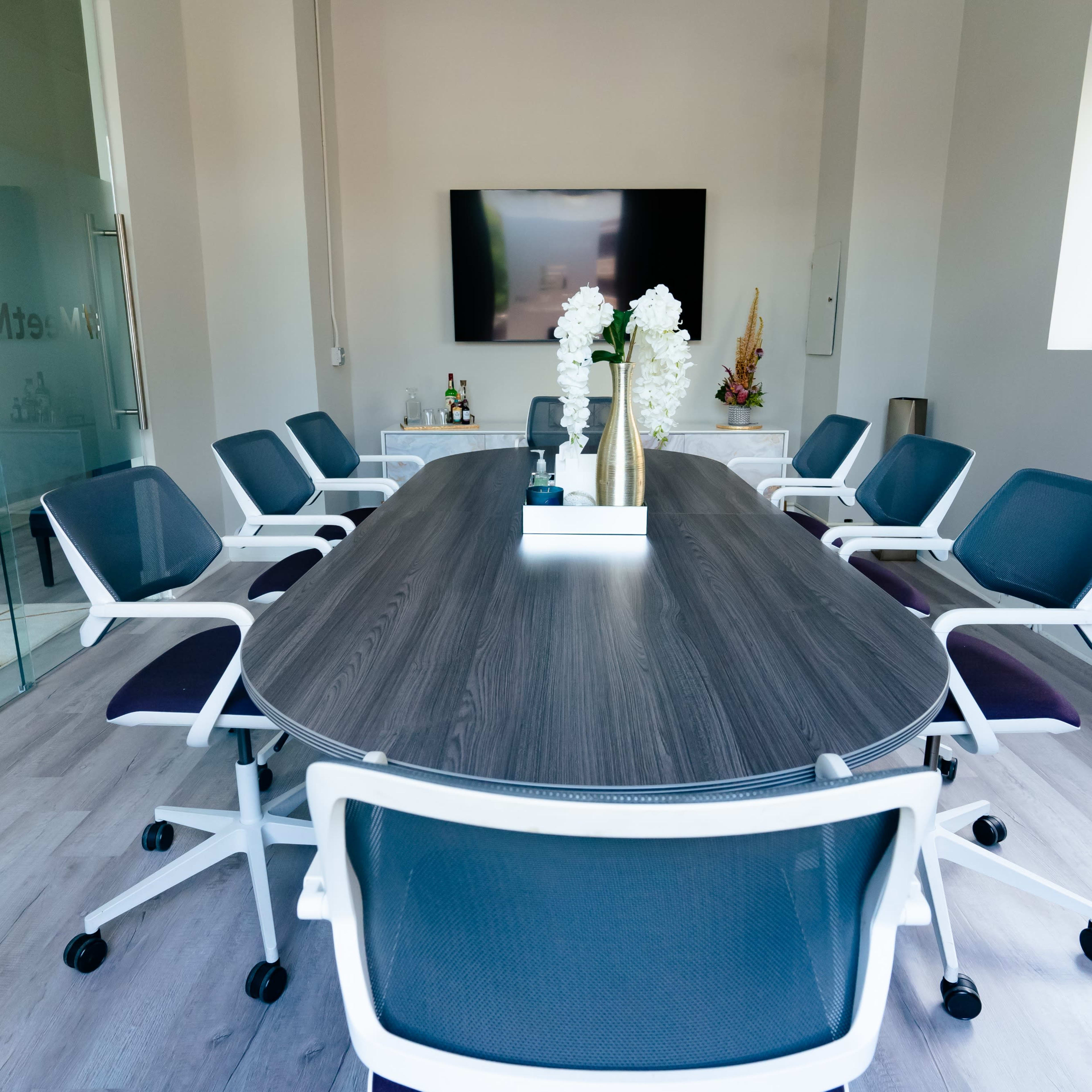 A modern conference room features a large oval table surrounded by white and gray rolling chairs, with a television mounted on the wall and a decorative centerpiece.