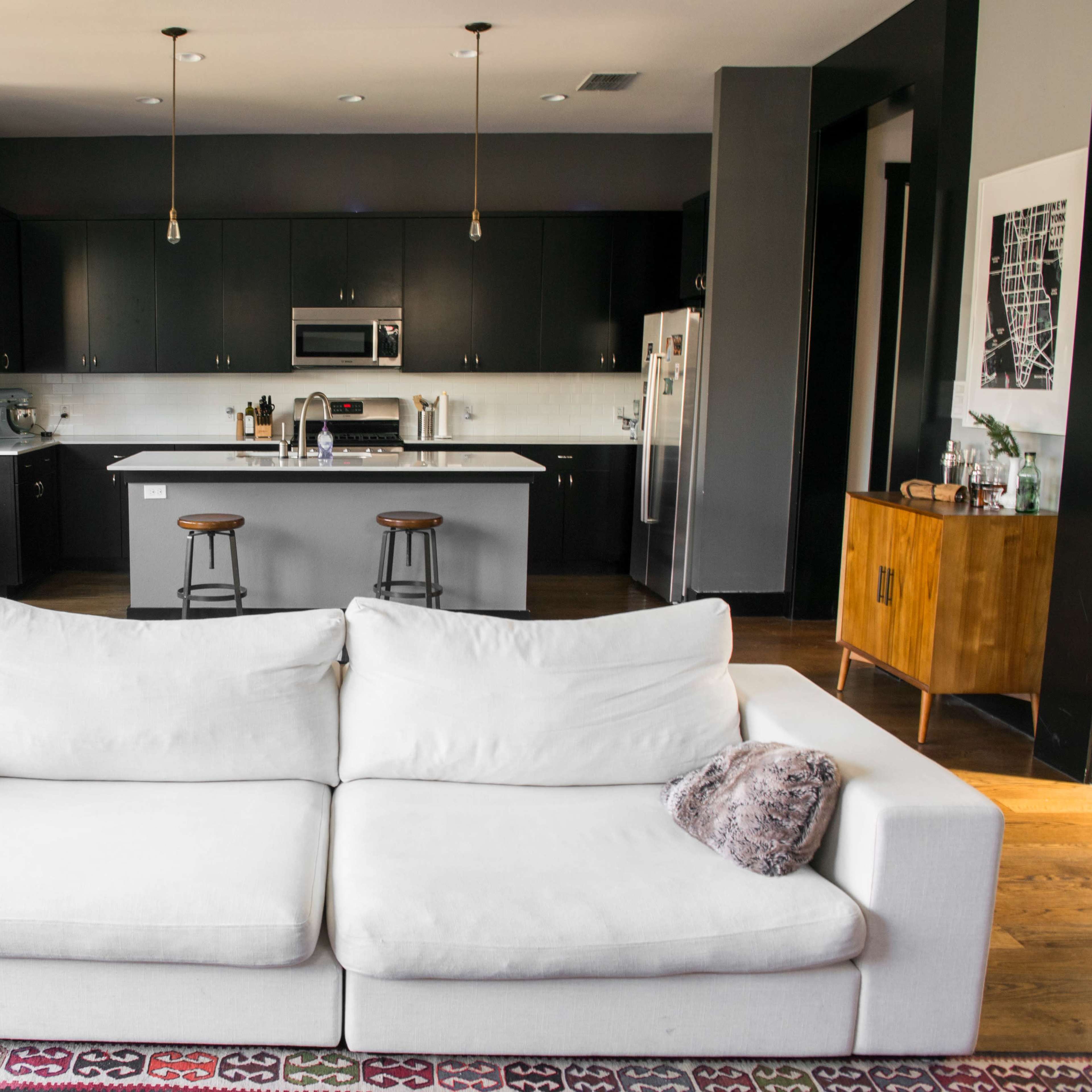 A modern living room features a white sofa facing a kitchen with dark cabinetry and a dining area in the background.