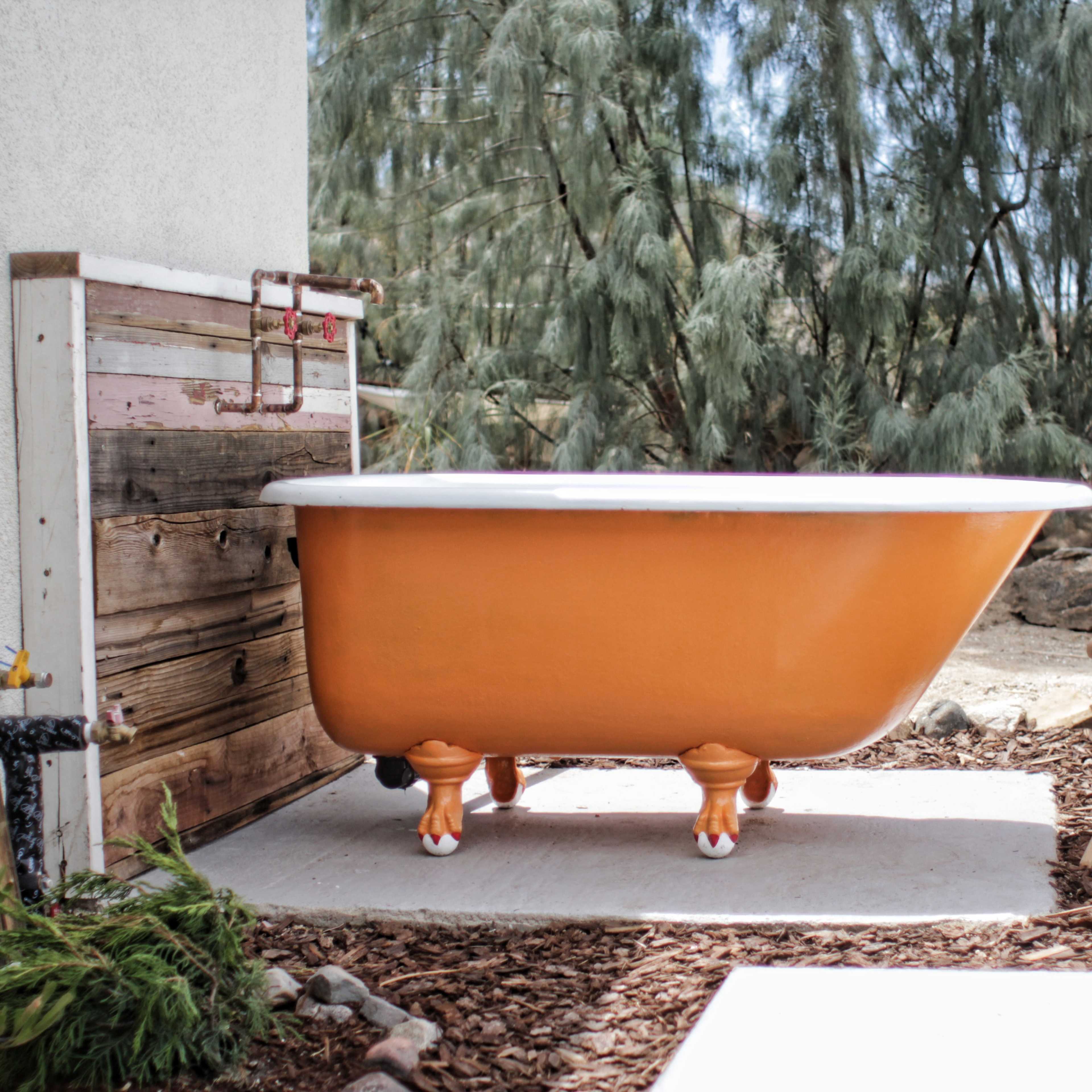 A bright orange clawfoot bathtub is placed outside on a stone pathway, surrounded by greenery and wood features.