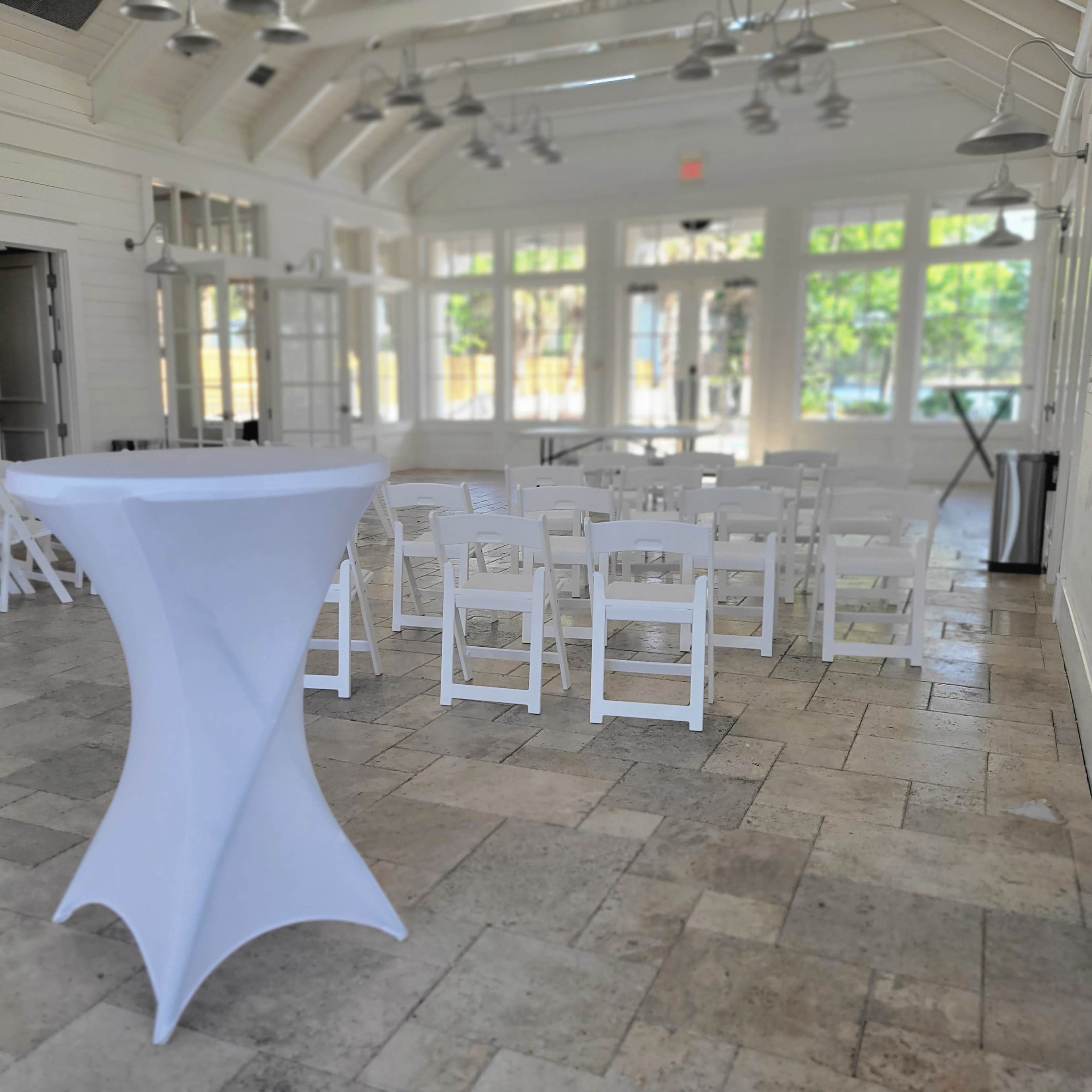 A brightly lit event space with white folding chairs arranged in rows facing a small stage, alongside a high-top table covered with a white tablecloth.