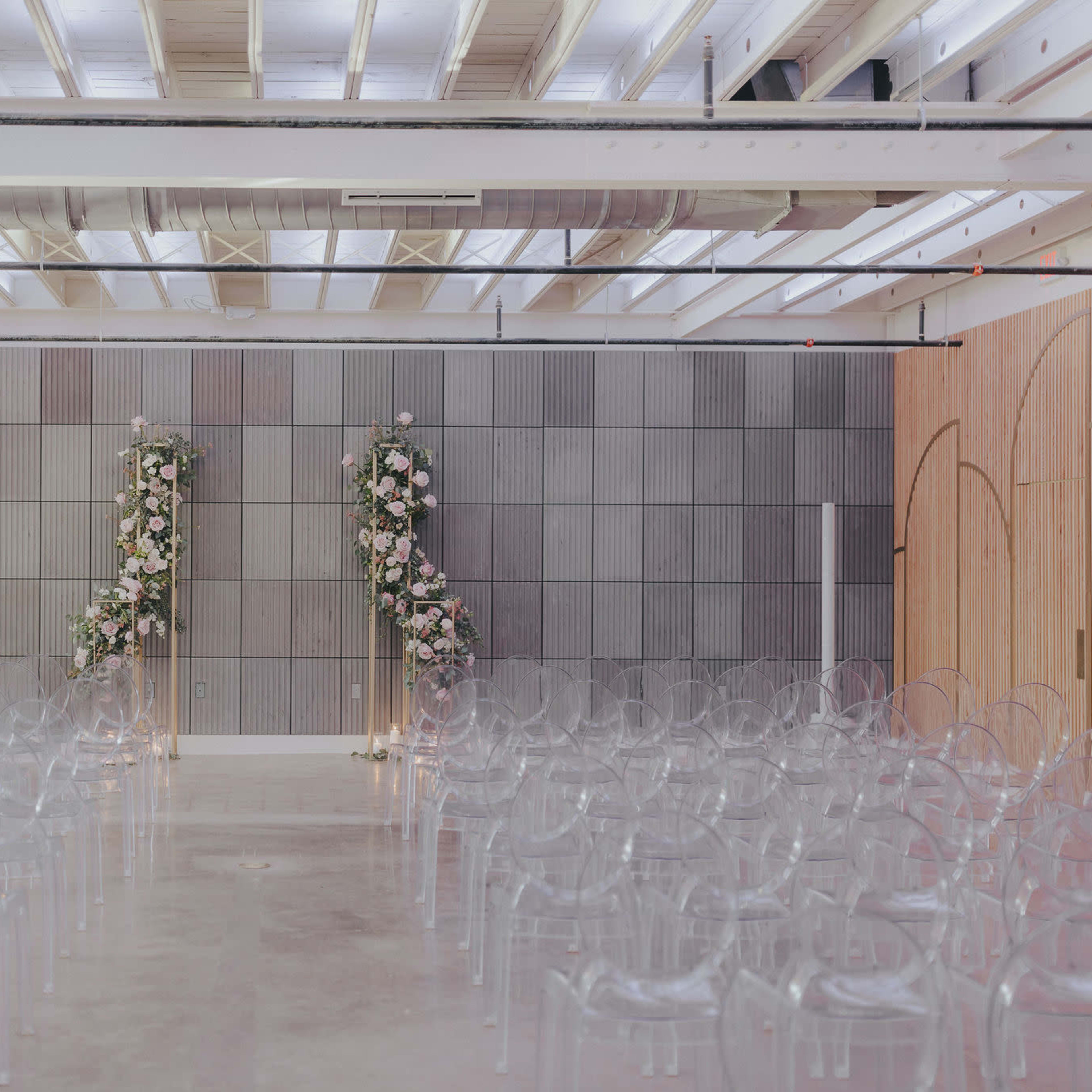 A modern wedding venue with transparent chairs arranged in rows facing a floral-adorned altar against a gray wall.