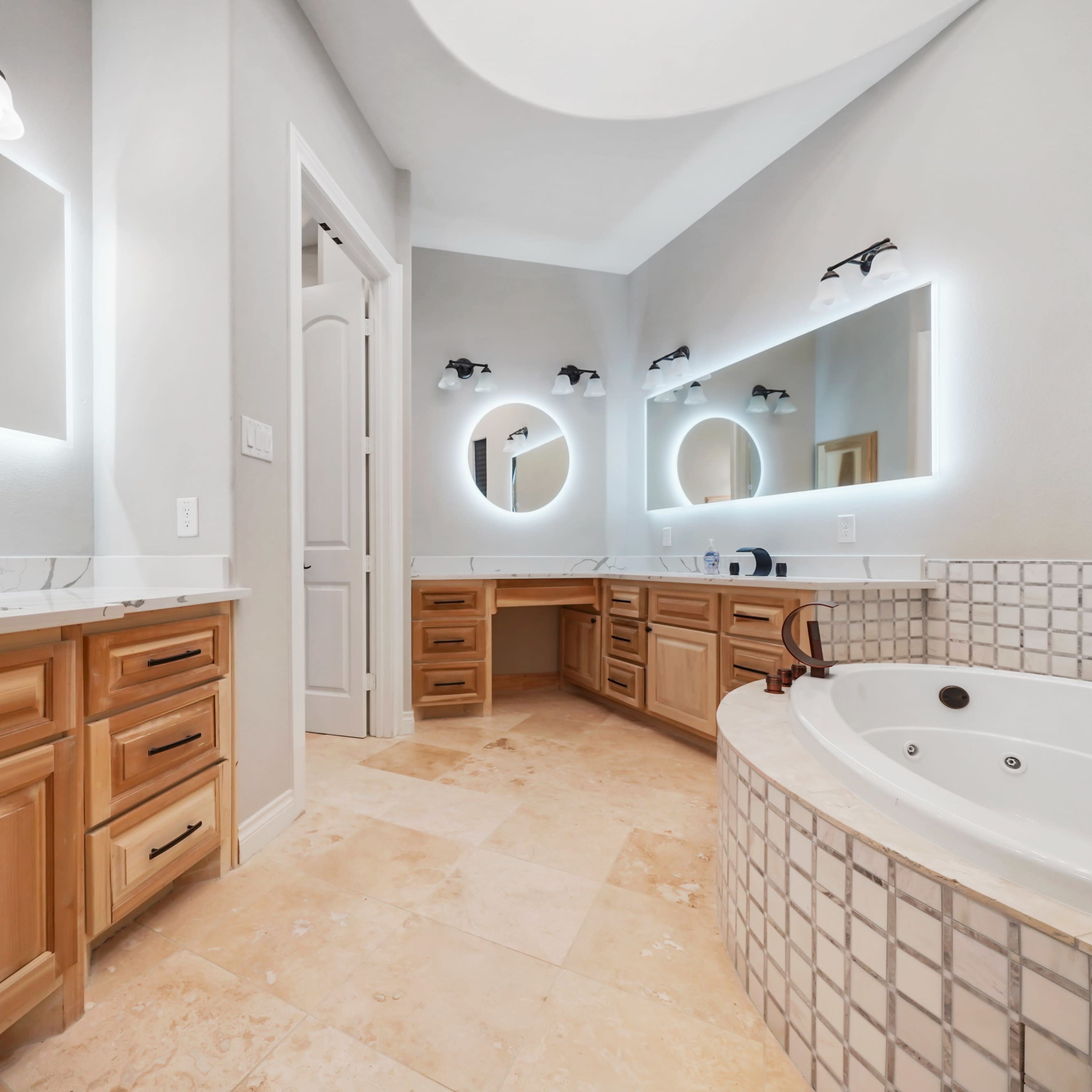 A spacious bathroom featuring dual sinks, a large circular mirror, wooden cabinetry, and a jetted bathtub.