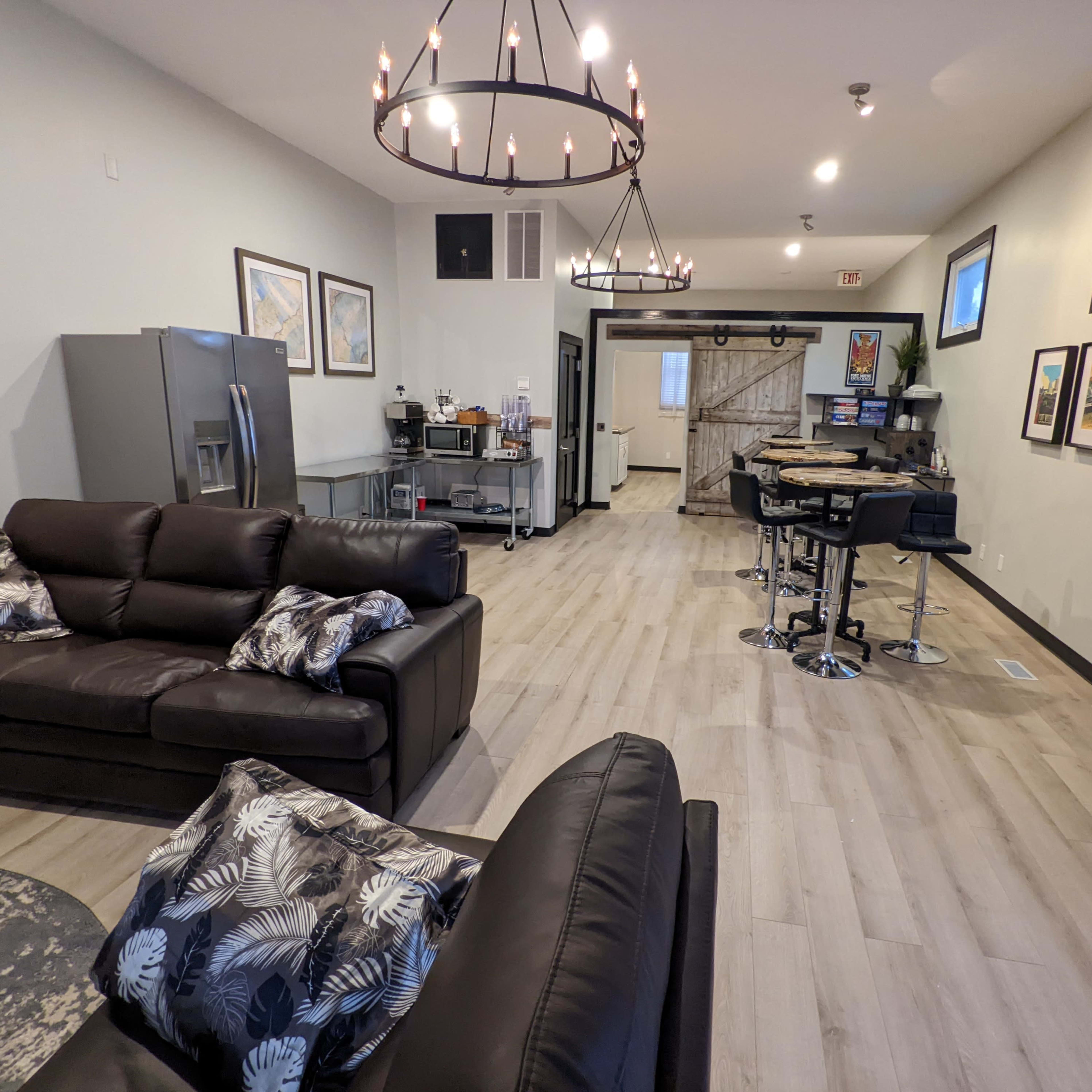 A modern, open living space featuring two black couches, a dining area with bar stools, and a kitchenette with stainless steel appliances.