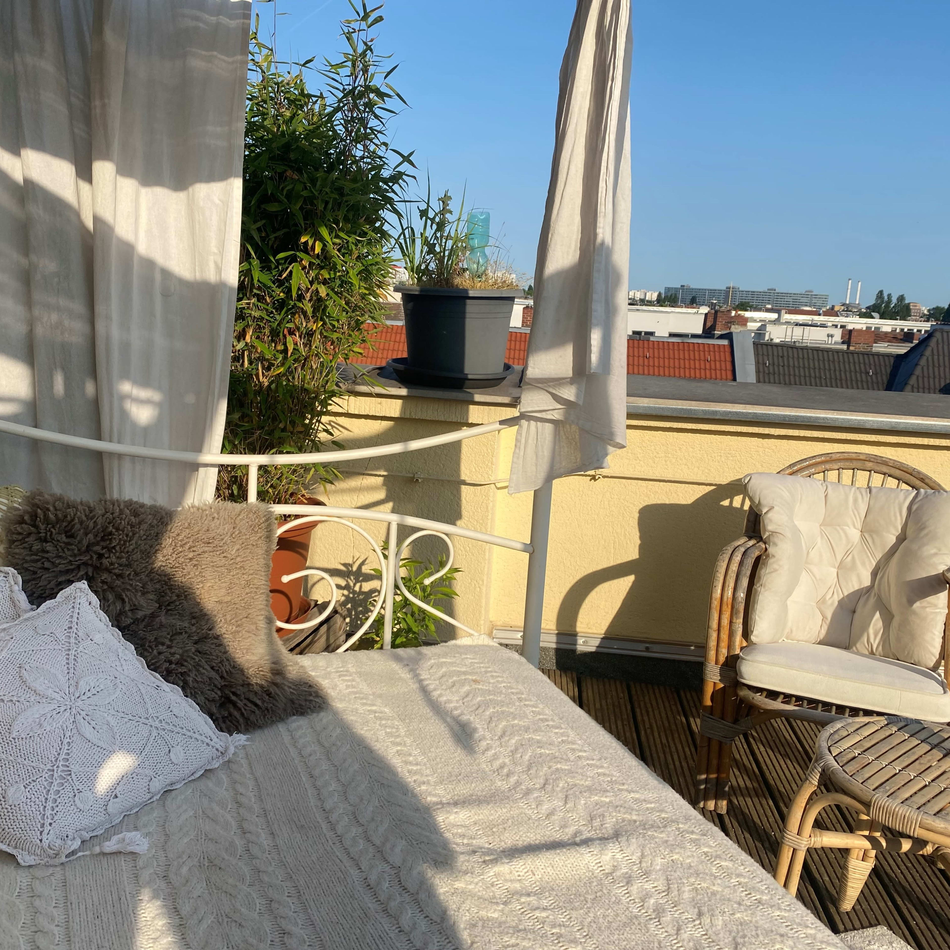A sunlit balcony features a white metal bed with pillows, a wicker chair, and potted plants against a clear blue sky.