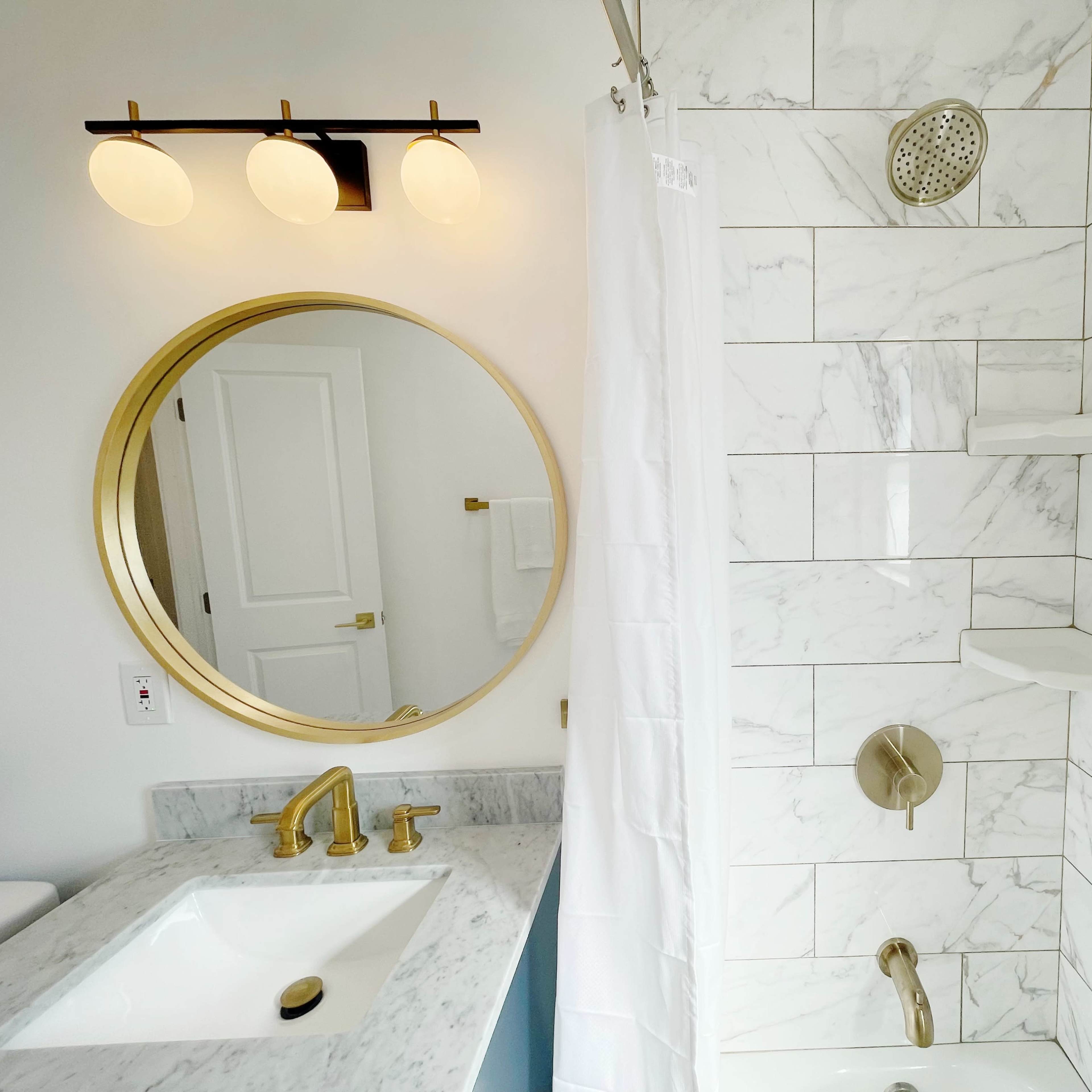 A modern bathroom with a marble-tiled shower, a round mirror, and a sink with gold fixtures.