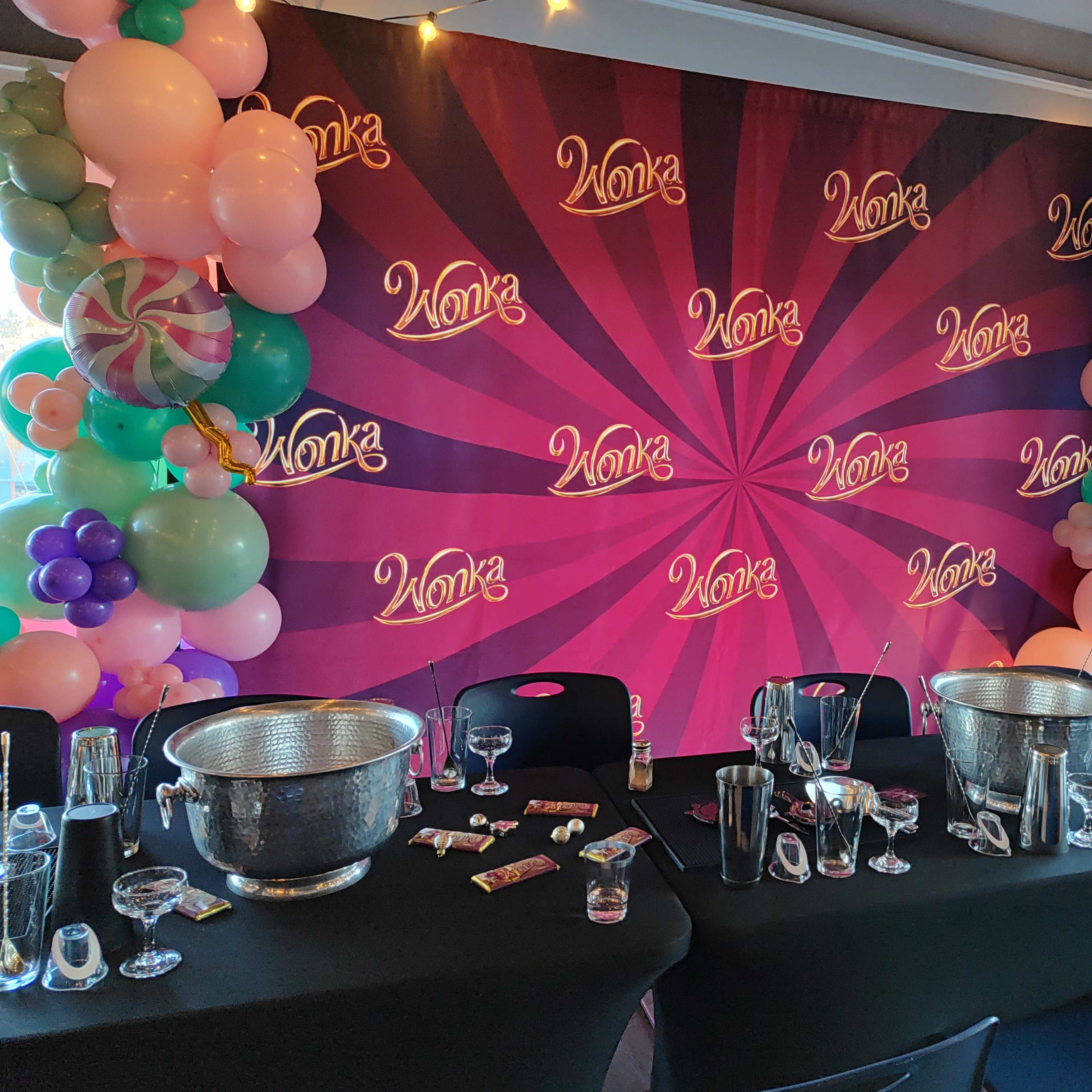A decorated table is set for a "Wonka" themed party, featuring a vibrant backdrop, colorful balloons, and glassware arranged neatly.