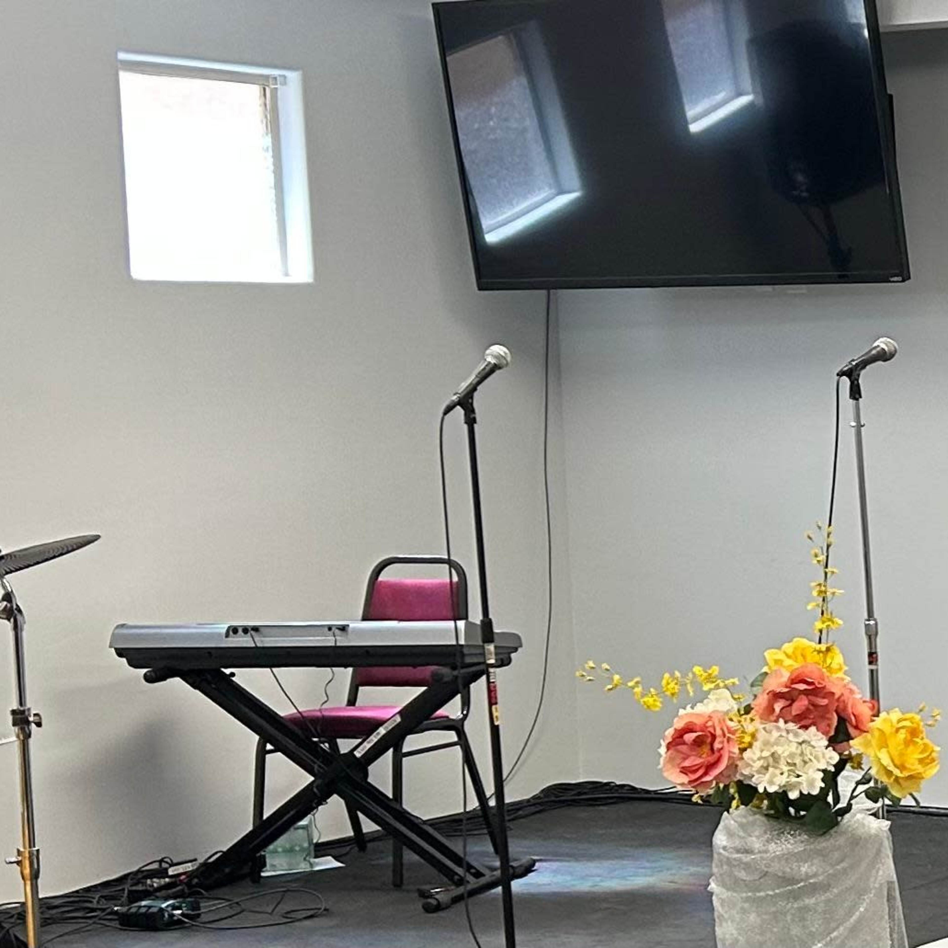 The scene features a musical setup with a drum kit, keyboard, two microphones, a TV screen, and a floral arrangement on a stage.