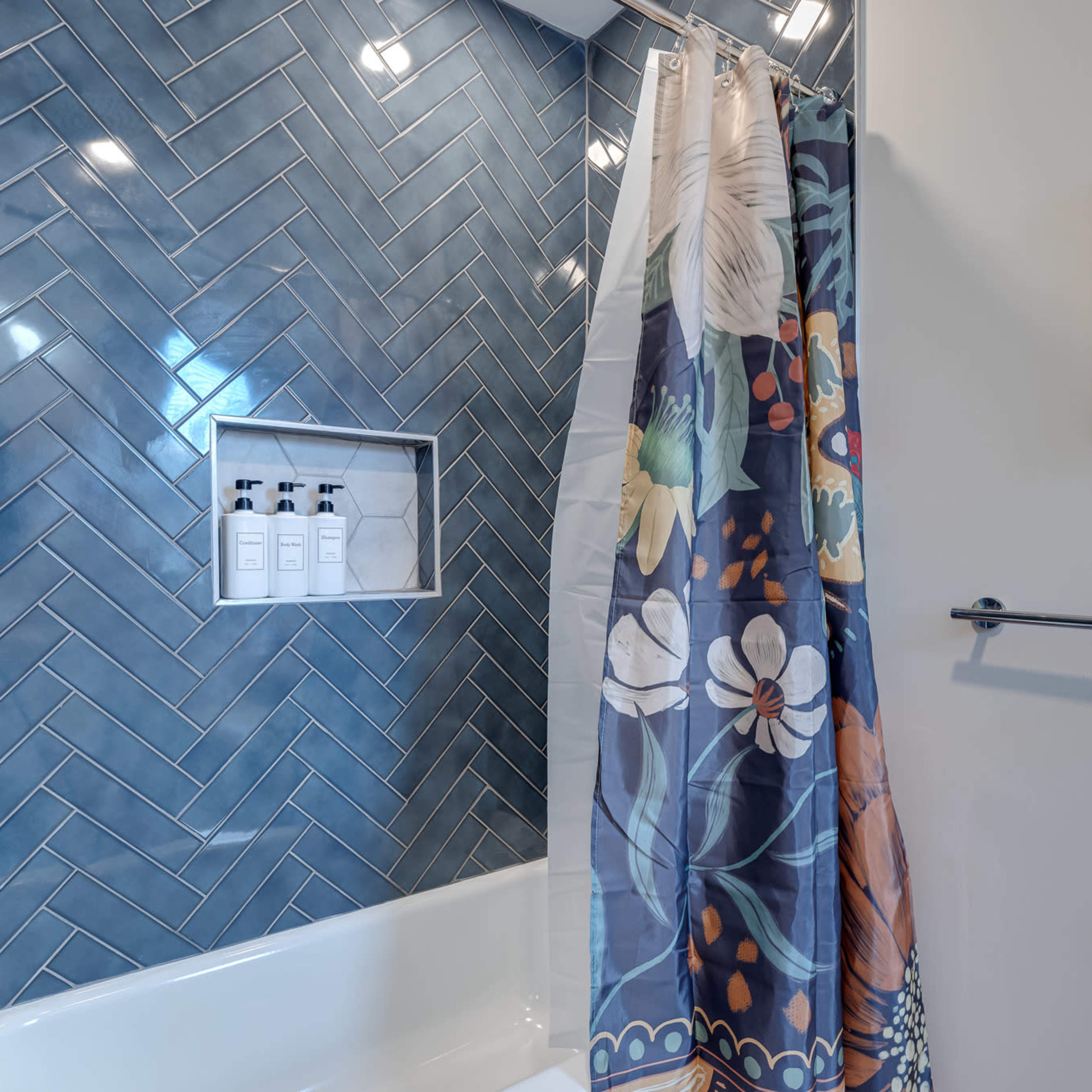 A bathroom with a blue herringbone tile wall, a white bathtub, and a floral shower curtain.