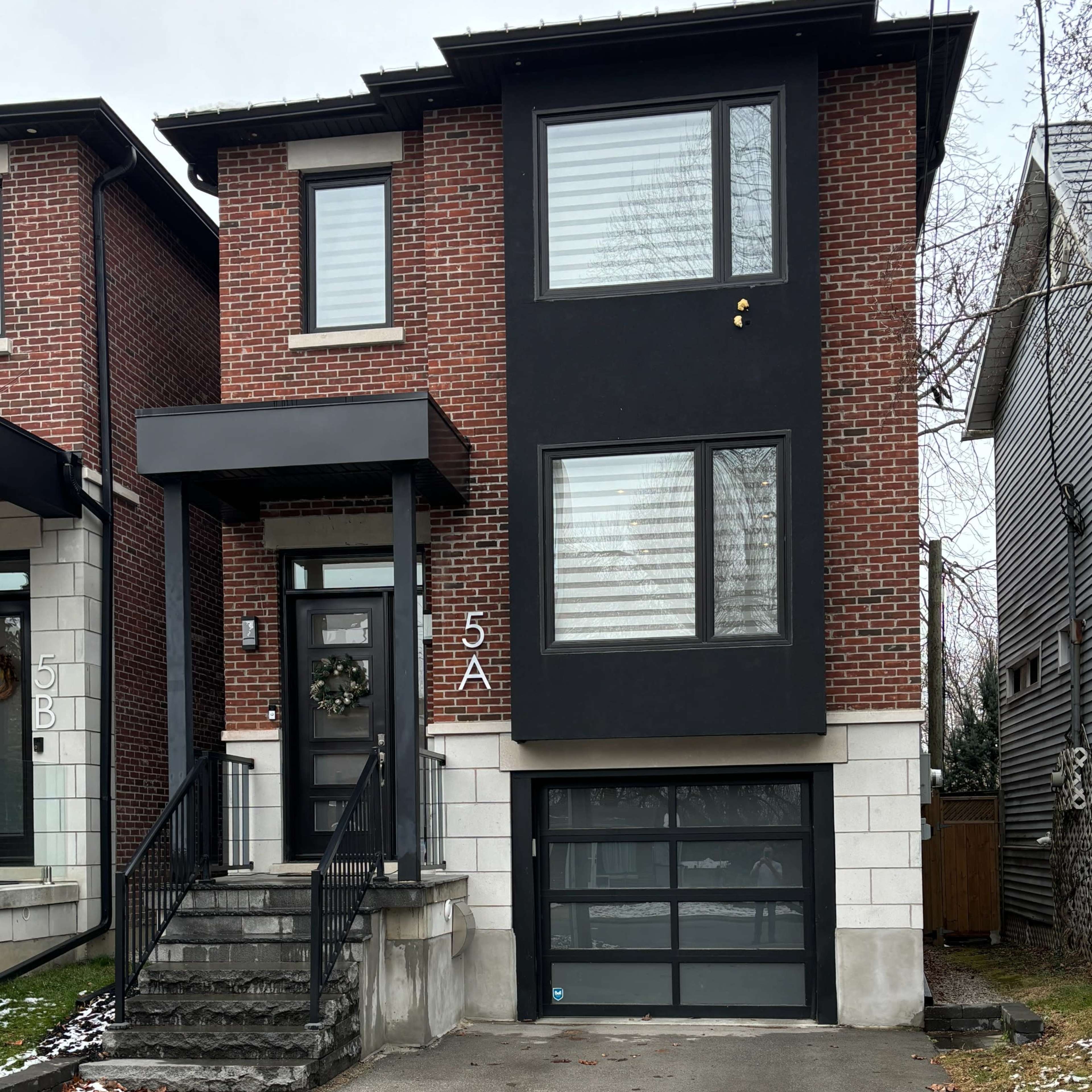 A modern two-story brick house with a black exterior and a garage door, set on a residential street with a partially snow-covered driveway.
