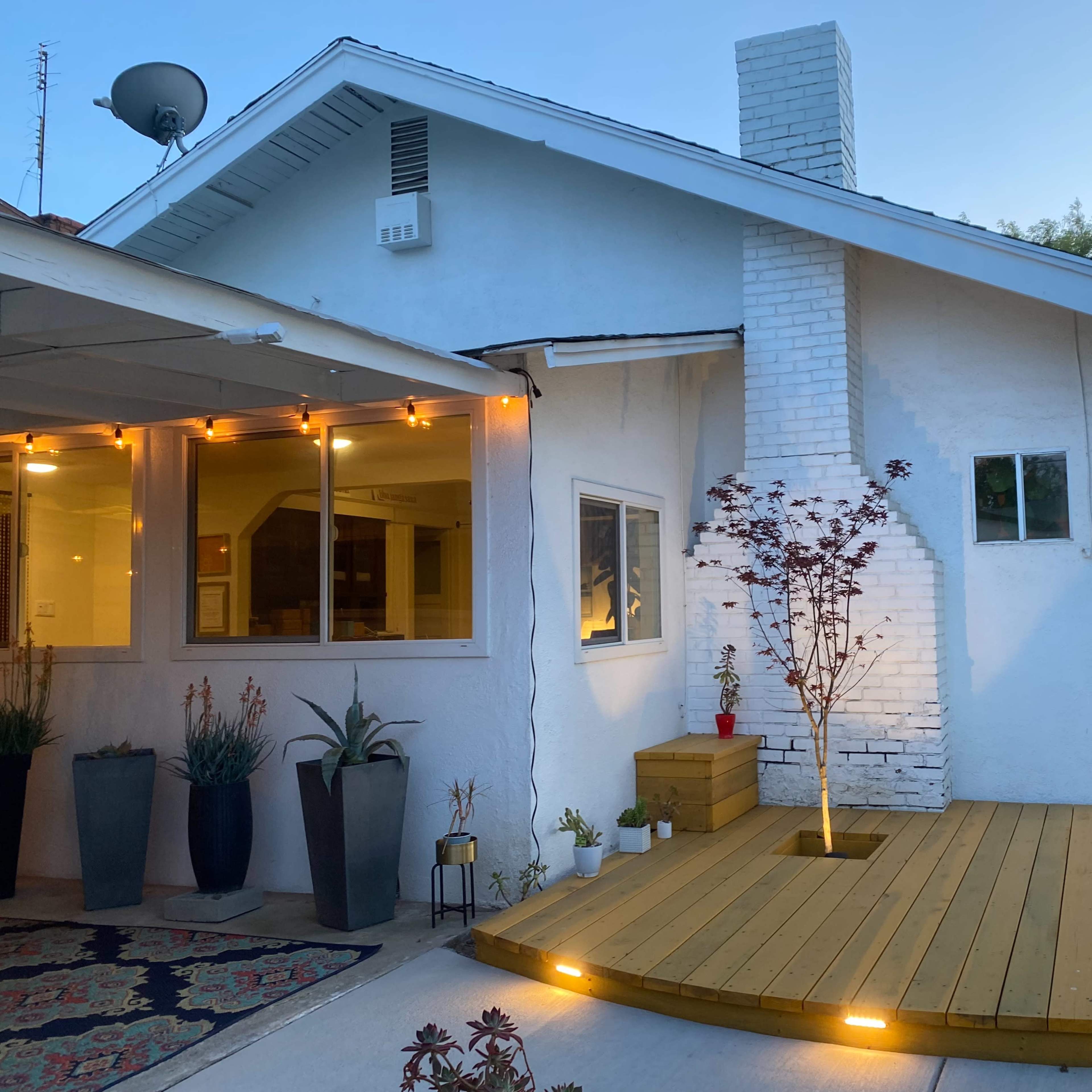 A residential house with a white brick chimney, a wooden deck, and outdoor lighting, situated at the corner of a street.