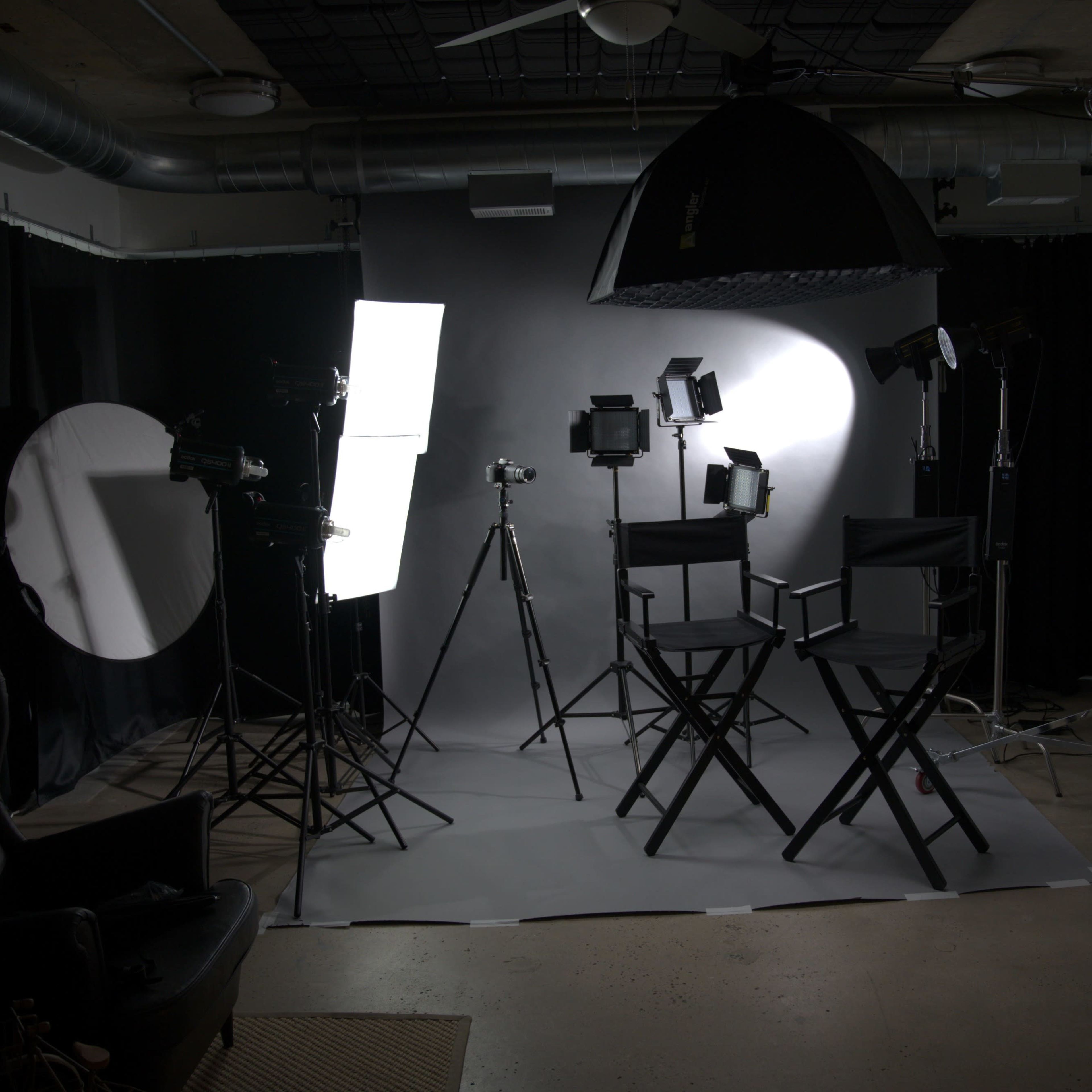 A photography studio is set up with multiple lights, cameras on tripods, and two empty director's chairs against a dark backdrop.