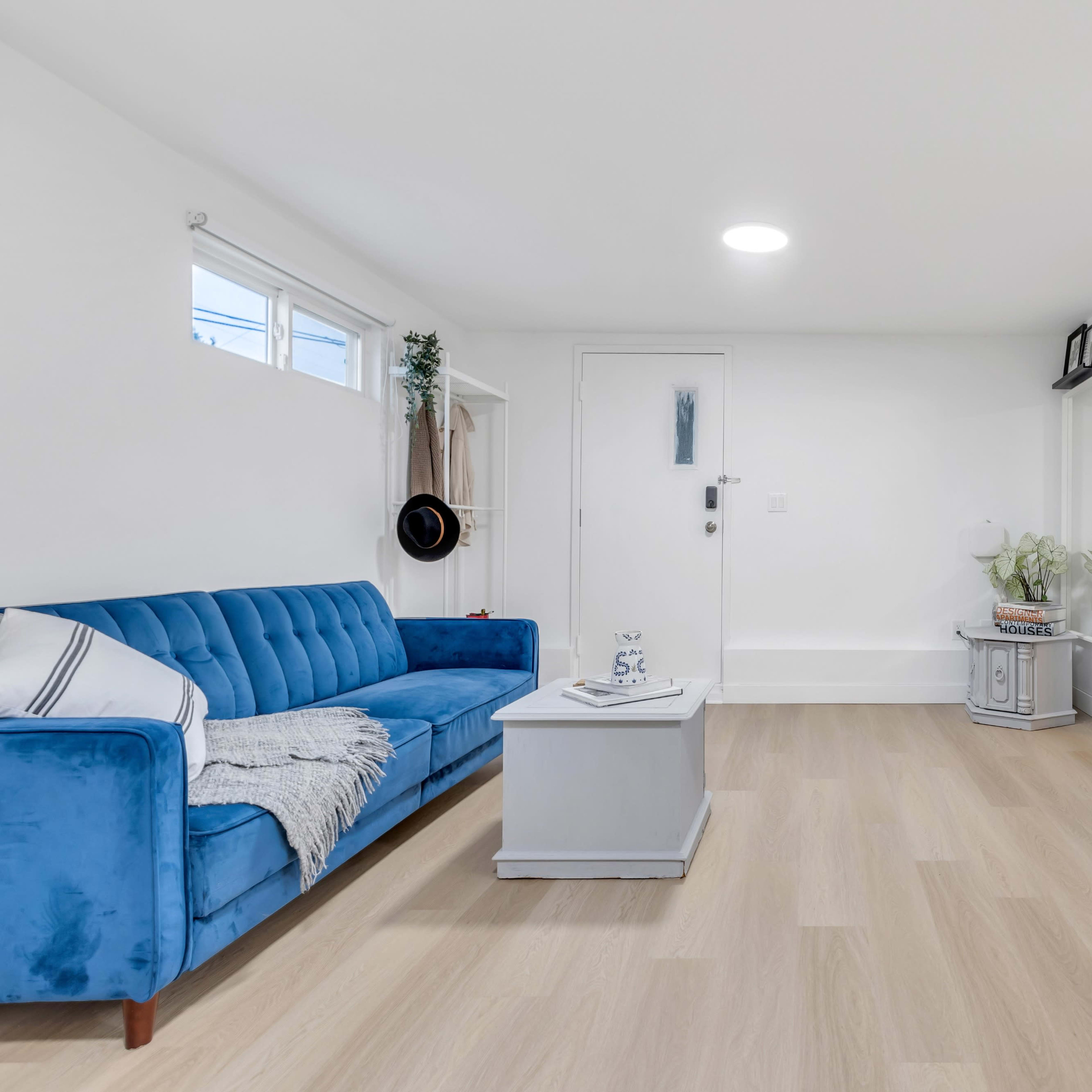 A minimalist living room featuring a blue couch, a light-colored coffee table, and a white console table against pale wood flooring.