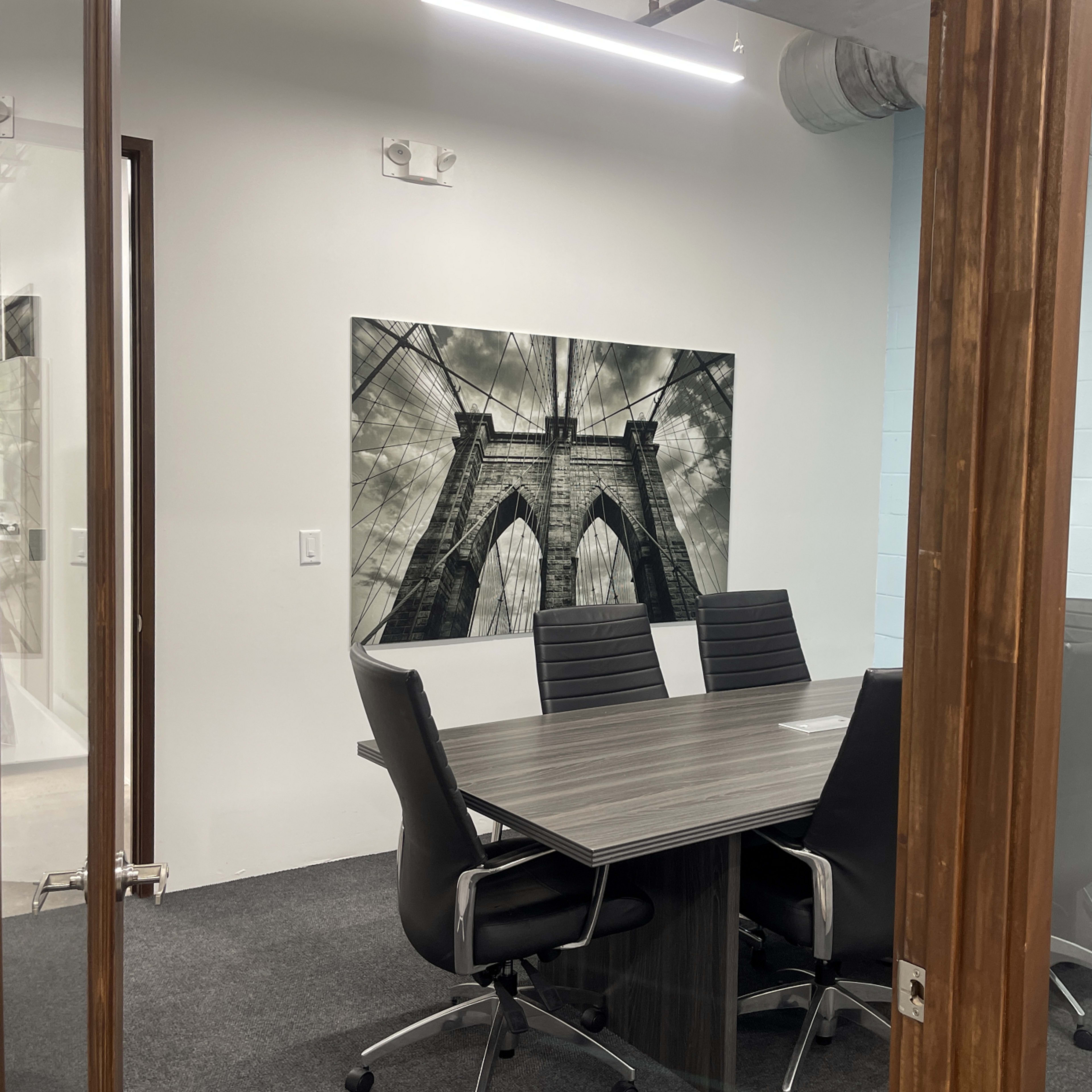 A glass-enclosed conference room features a large photograph of the Brooklyn Bridge on the wall, with a wooden table and black chairs arranged around it.