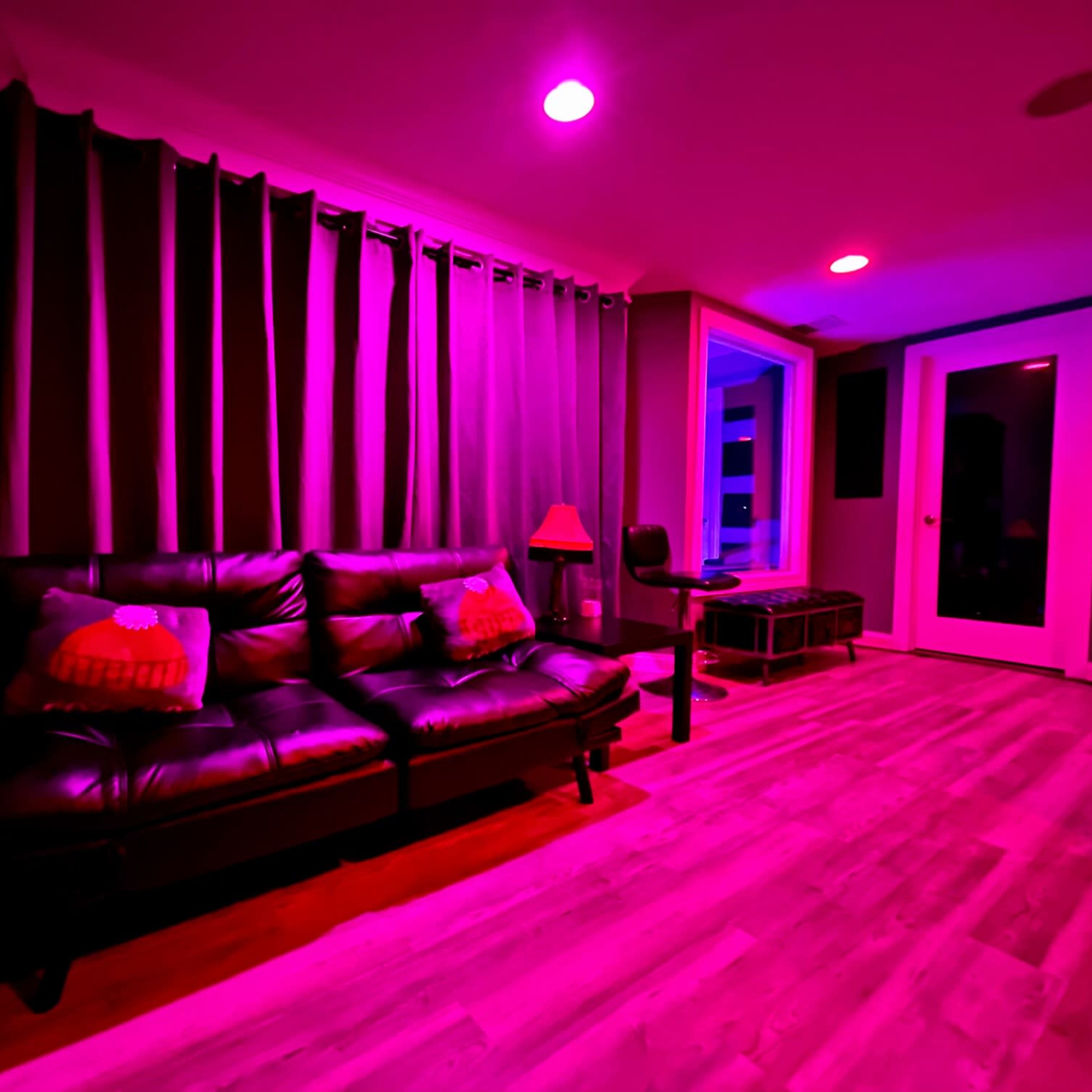 A modern living room features a black couch with pillows, a small lamp, and purple lighting, alongside curtains and large windows.