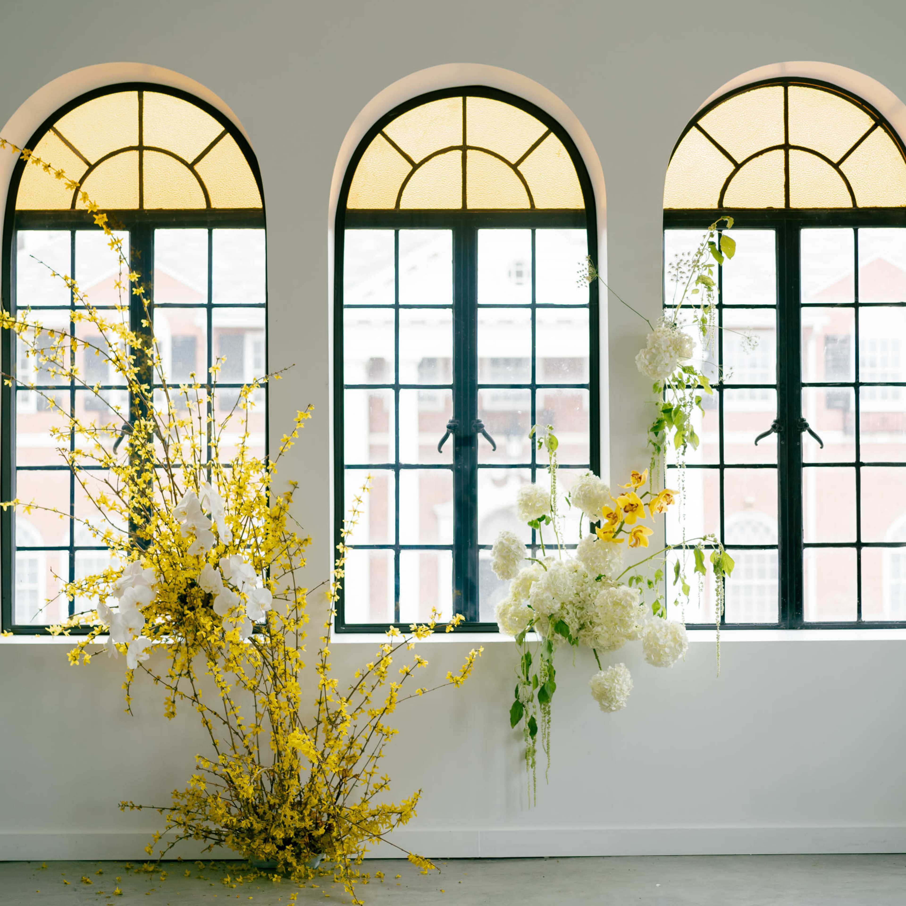 A minimalist interior with large arched windows and floral arrangements in yellow and white against a light-colored wall.