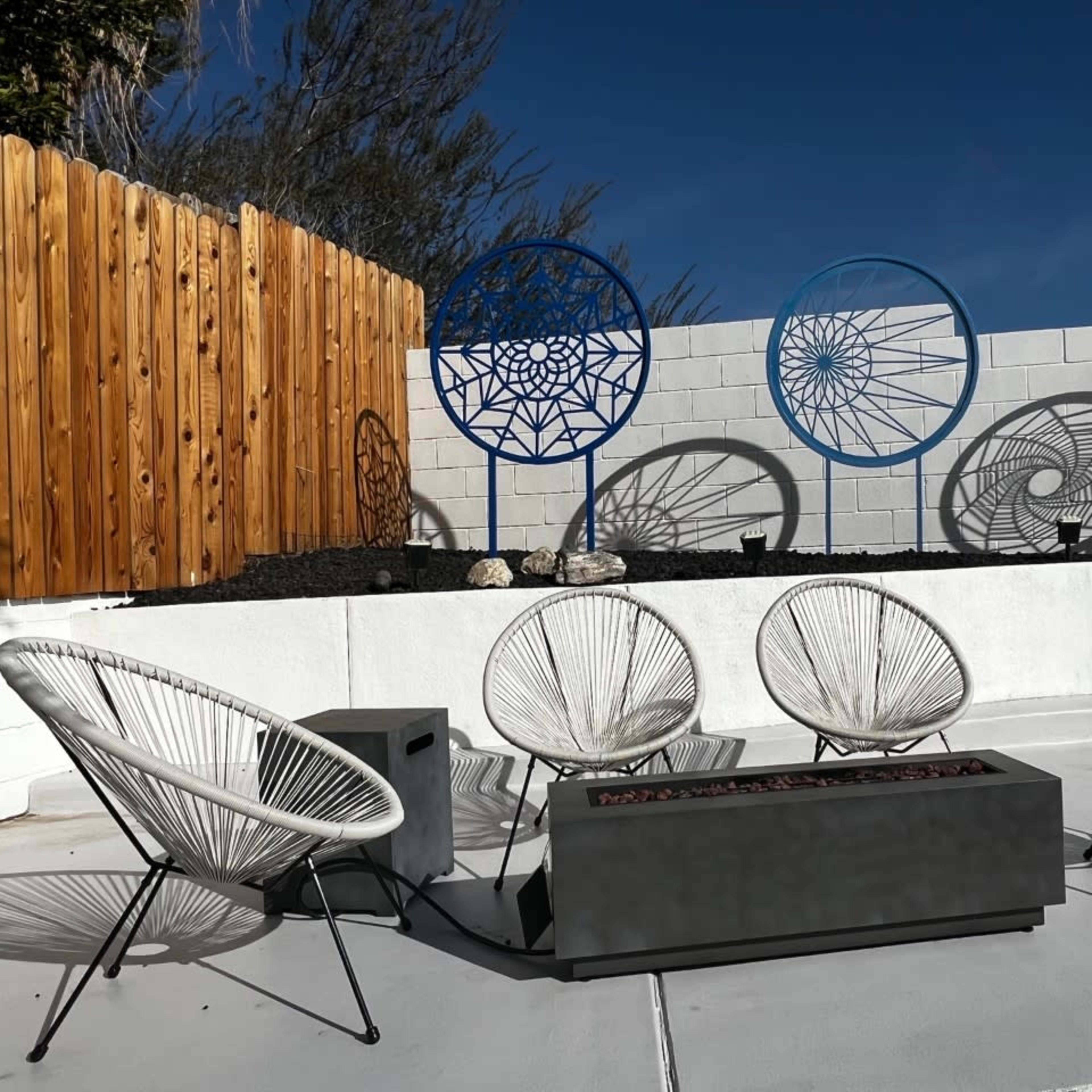 A patio area with five Acapulco chairs arranged around a rectangular fire pit, set against a white wall featuring decorative geometric wall art.