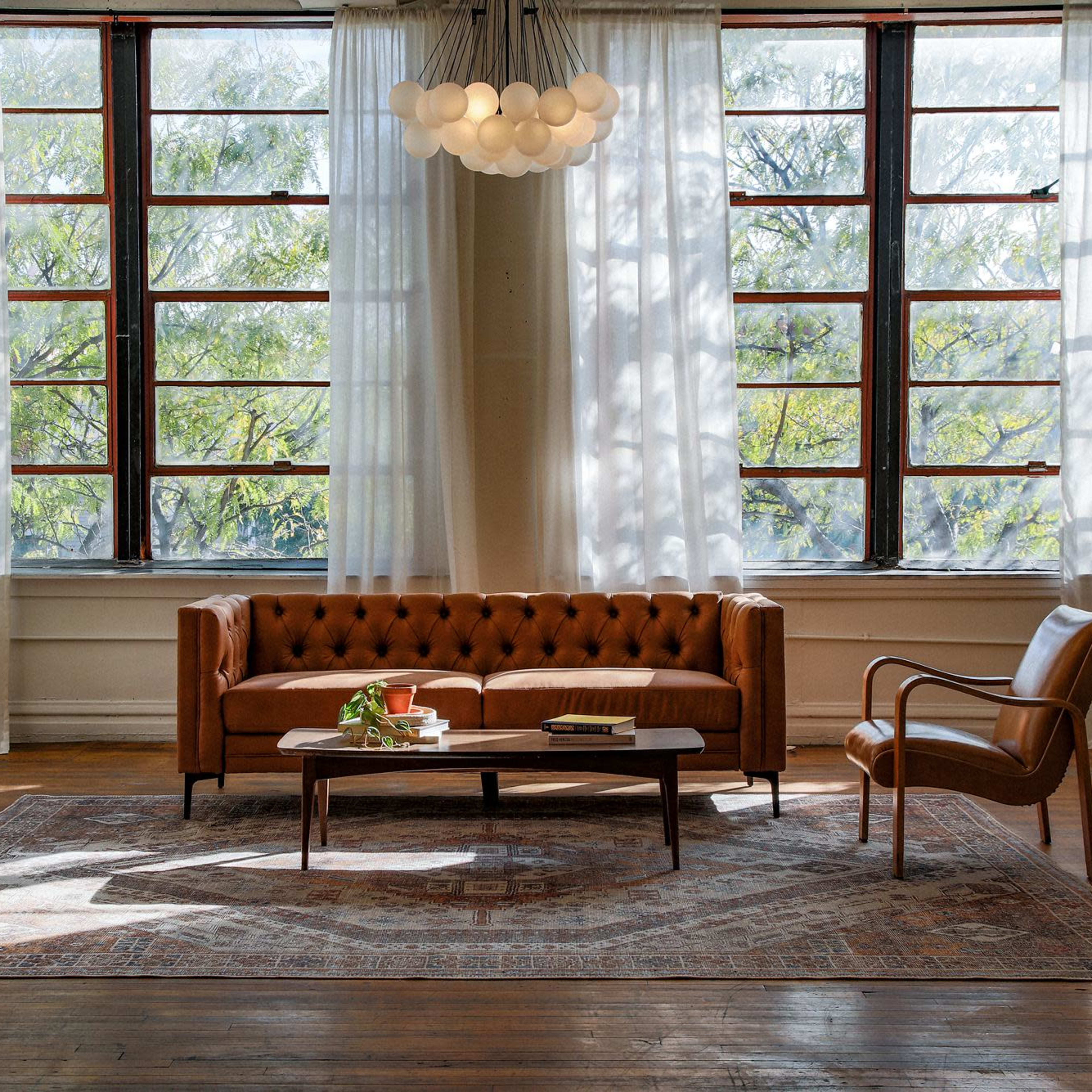 A sunlit living room featuring a brown leather couch, a wooden coffee table, and a single armchair, with large windows allowing greenery to be visible outside.