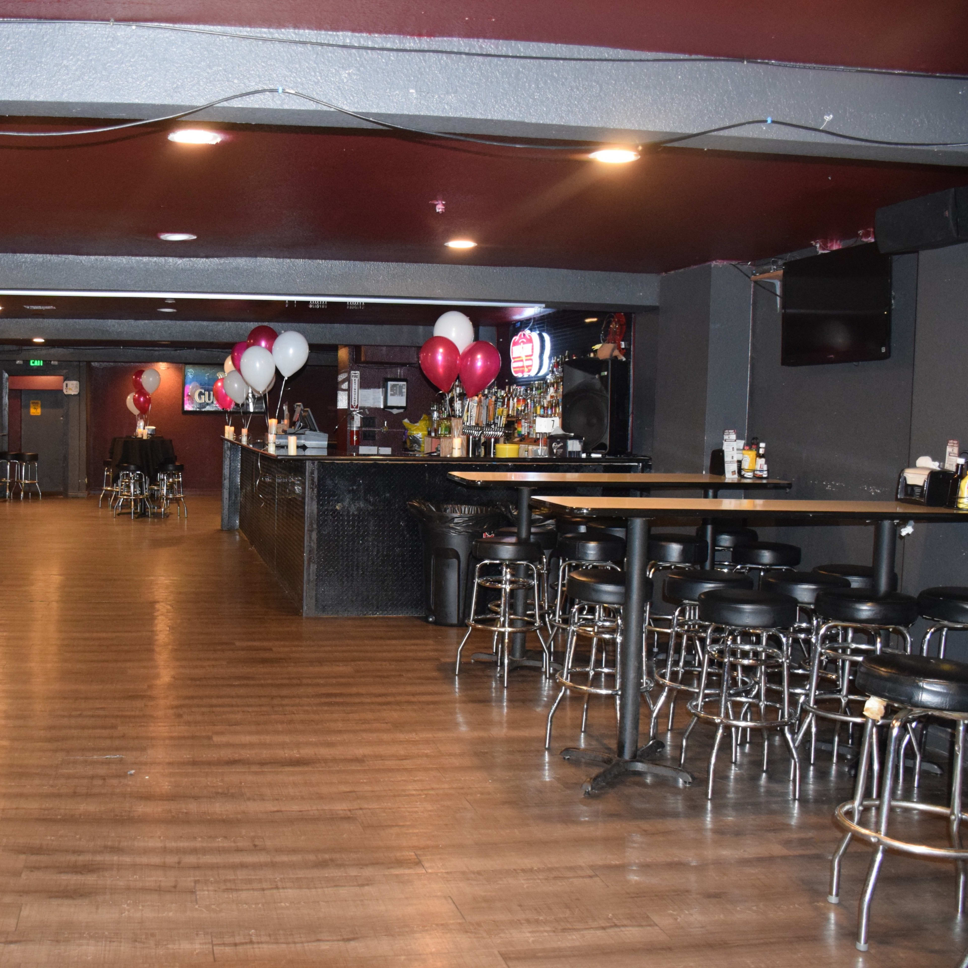 A spacious bar area with black walls, wooden flooring, several tables, chairs, and balloons arranged at one end.