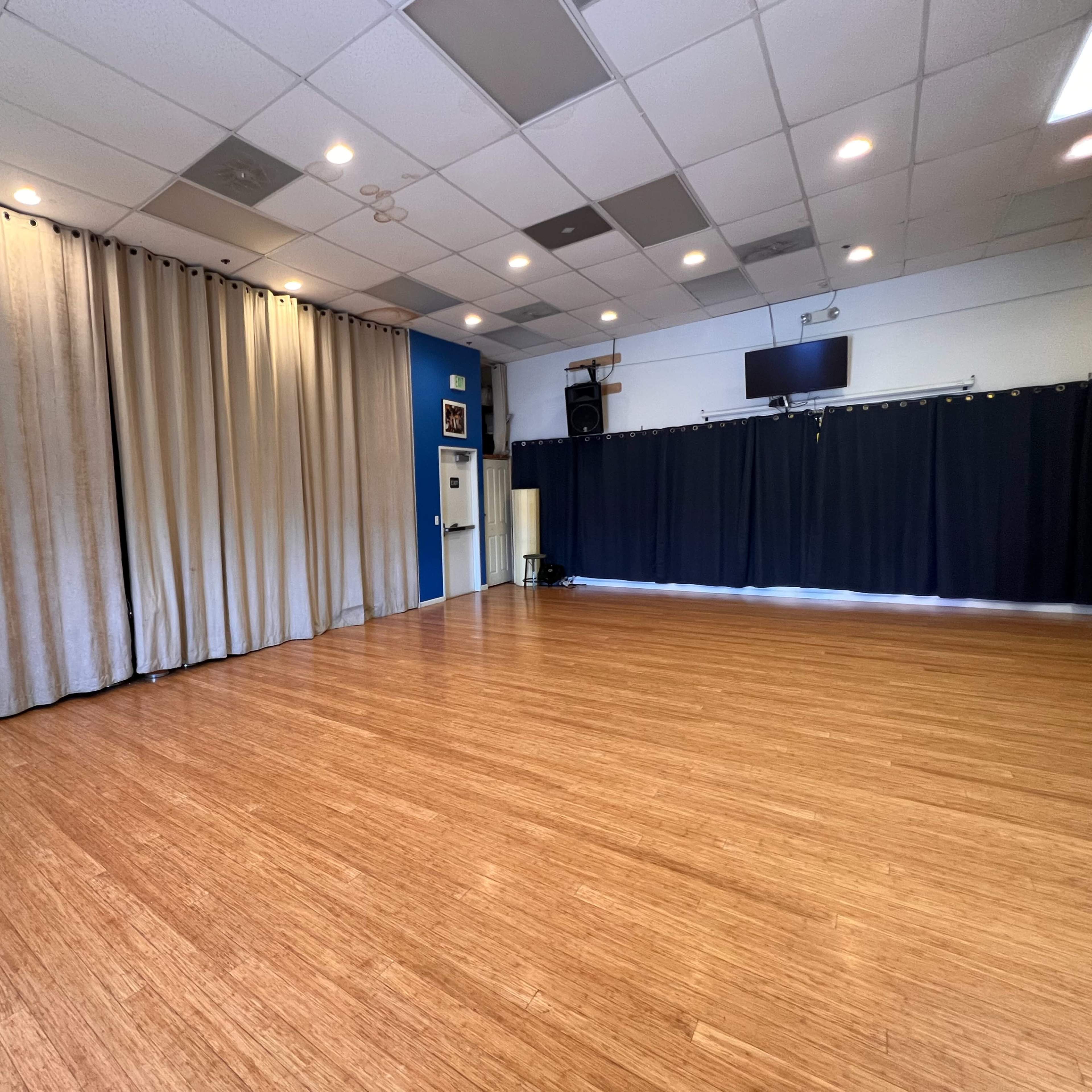 A spacious room with a wooden floor, dark curtains on one side, and beige curtains on the opposite side, along with wall-mounted equipment and lights.