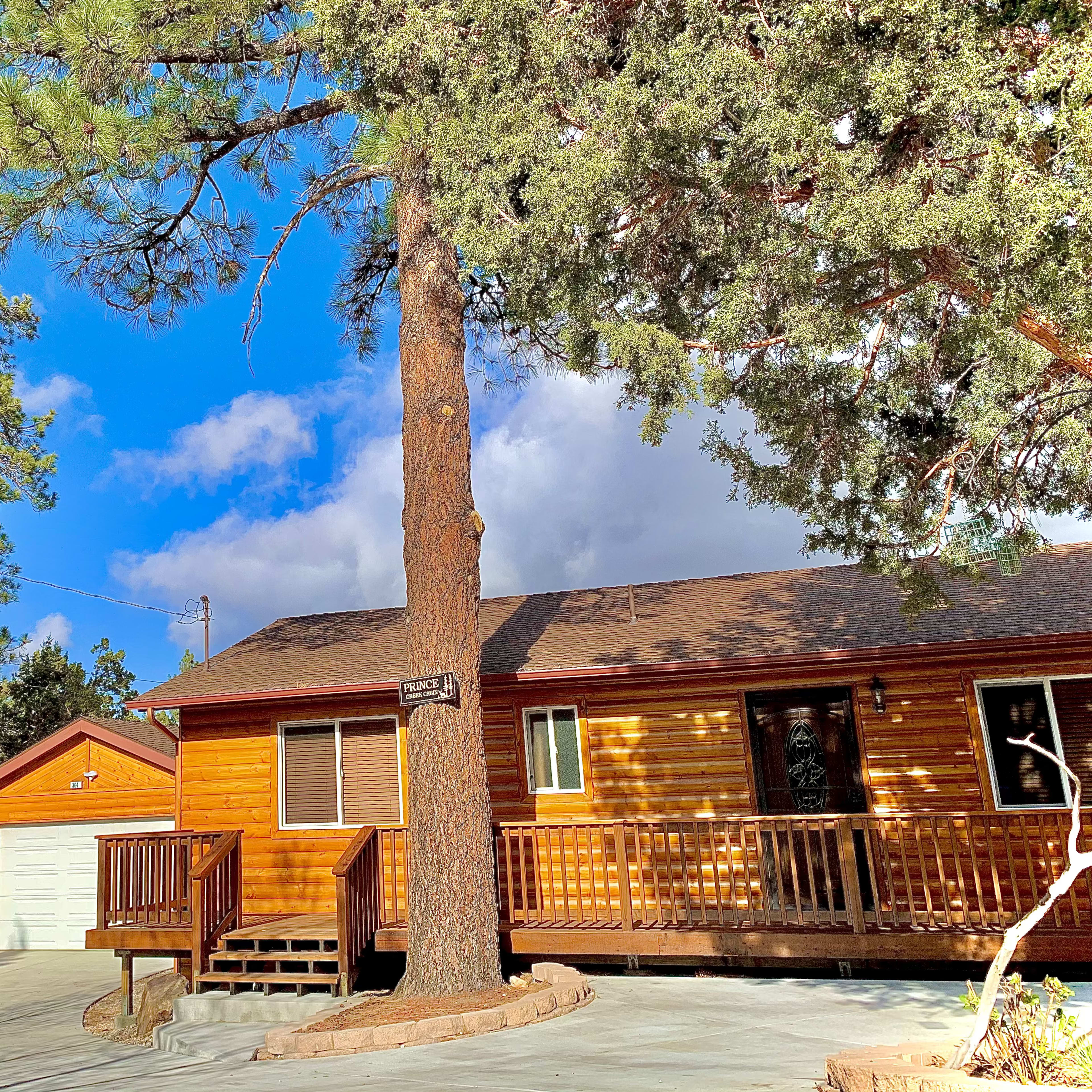A brown wooden cabin with a porch and a garage is surrounded by tall green trees under a partly cloudy sky.