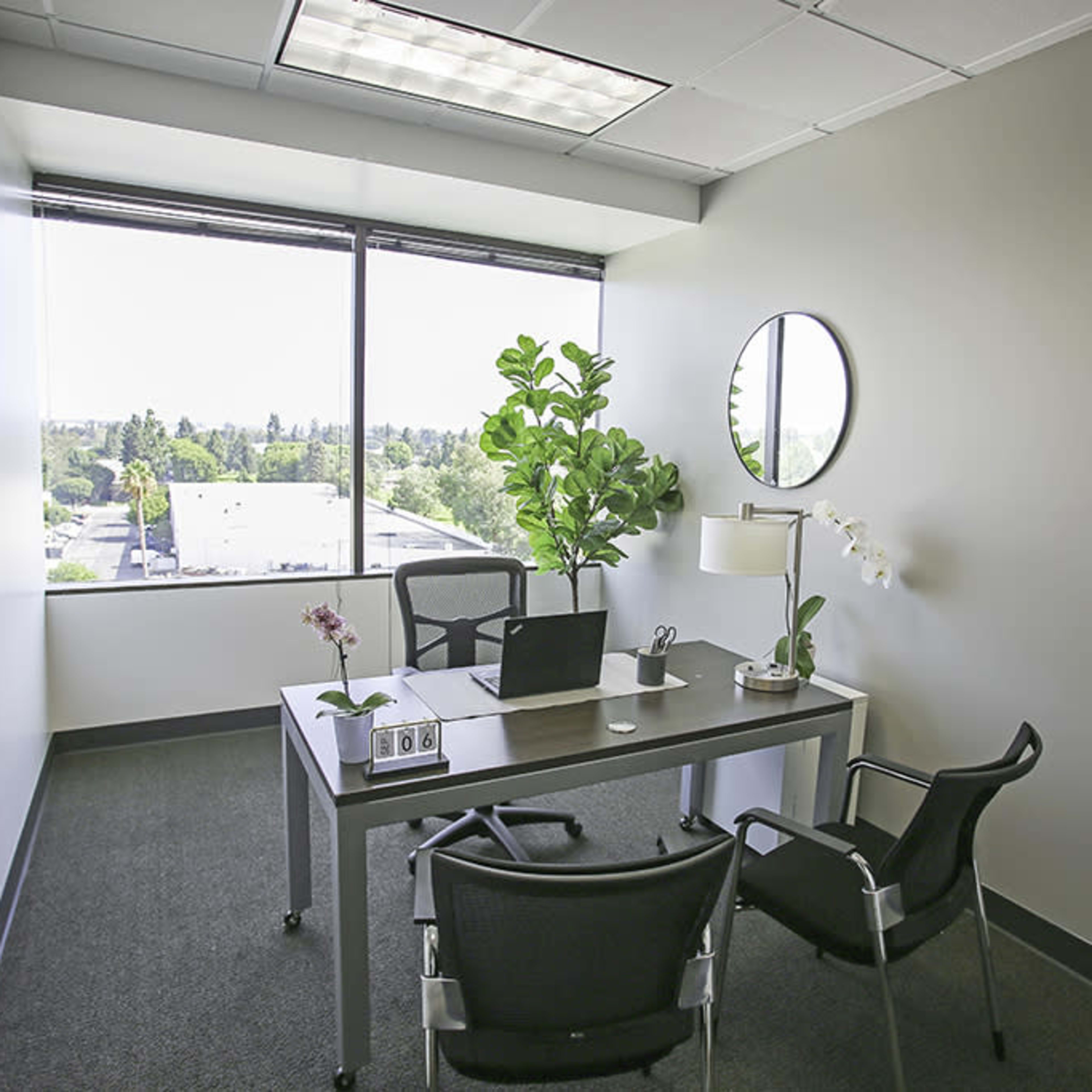 A small office features a desk with a laptop, a lamp, and a plant, accompanied by two chairs and a large window overlooking a landscape.