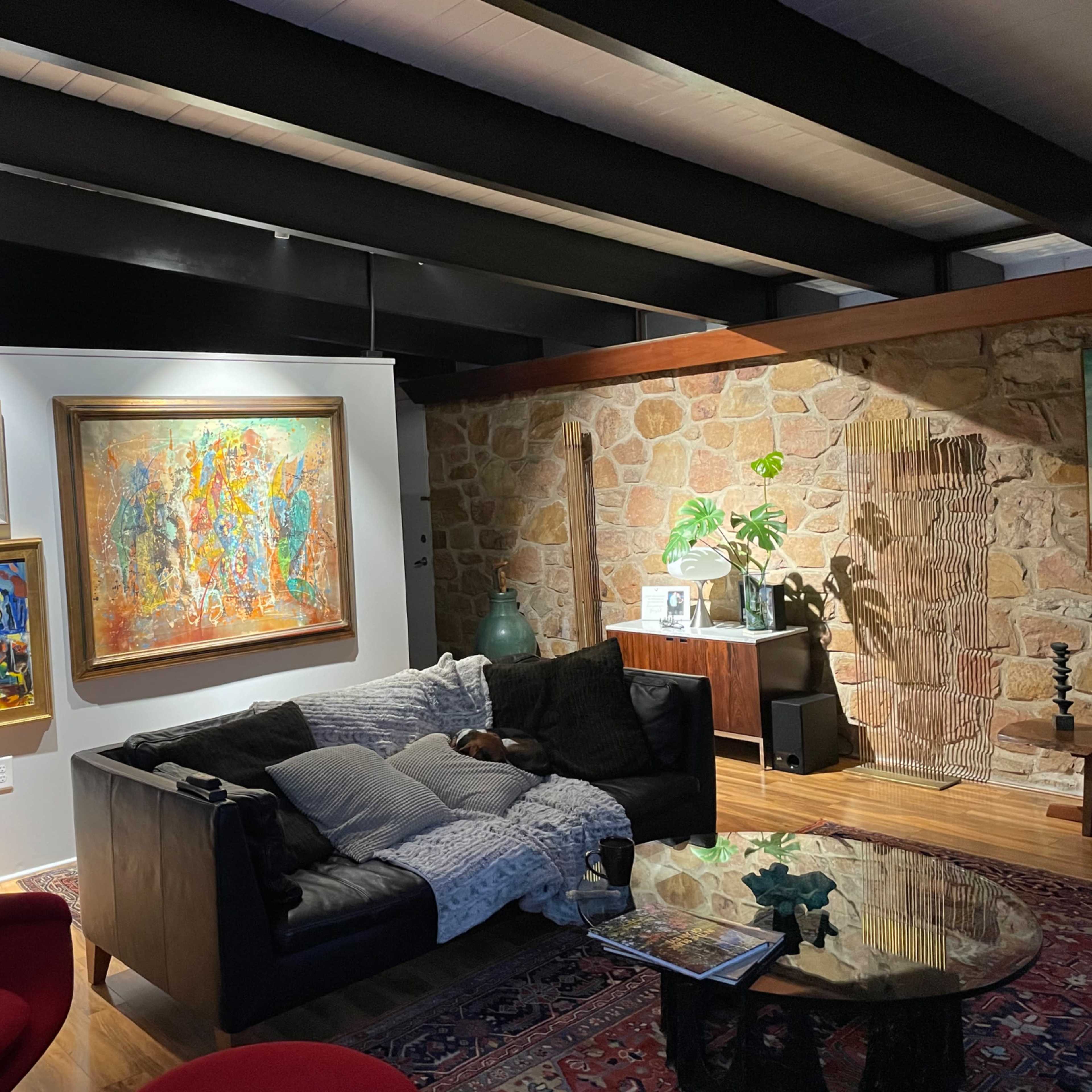 A living room features a black couch with a throw blanket, a round glass coffee table, colorful paintings on the wall, and a stone accent wall.